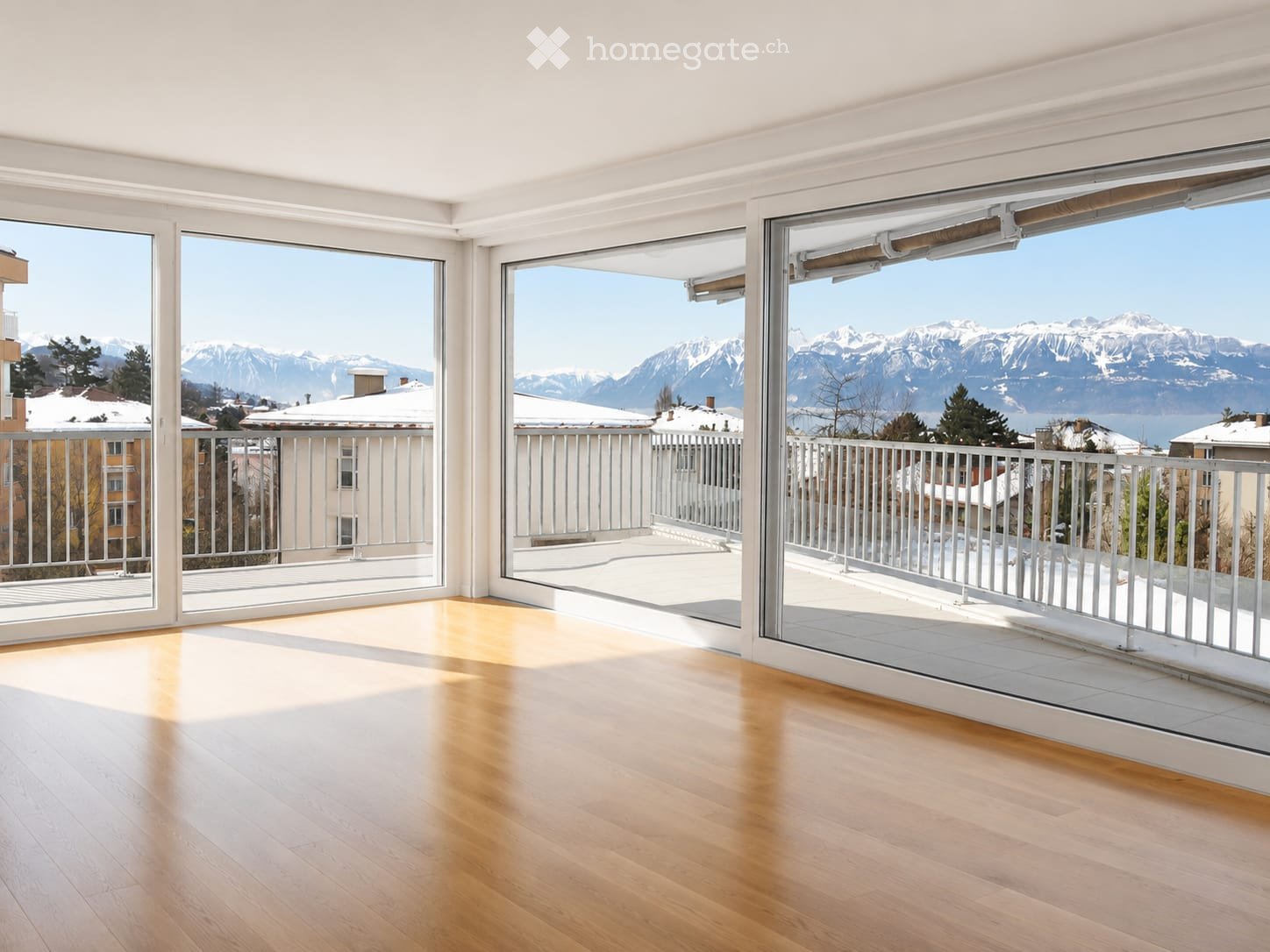 Bright, empty room with wooden flooring and large glass doors leading to a balcony with a scenic mountain view.