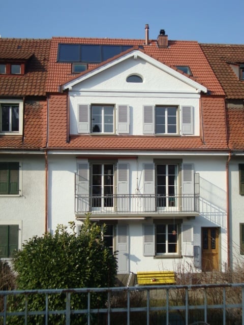 Two-story house with a red-tiled roof, white exterior, multiple windows with shutters, and a small balcony on the first floor.