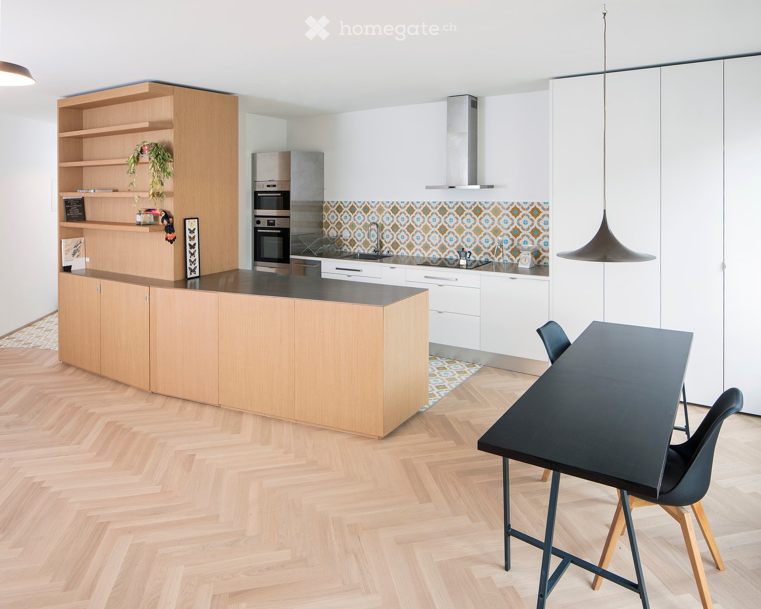 Modern kitchen with light wooden cabinets, a central island, stainless steel appliances, and a black dining table with chairs.
