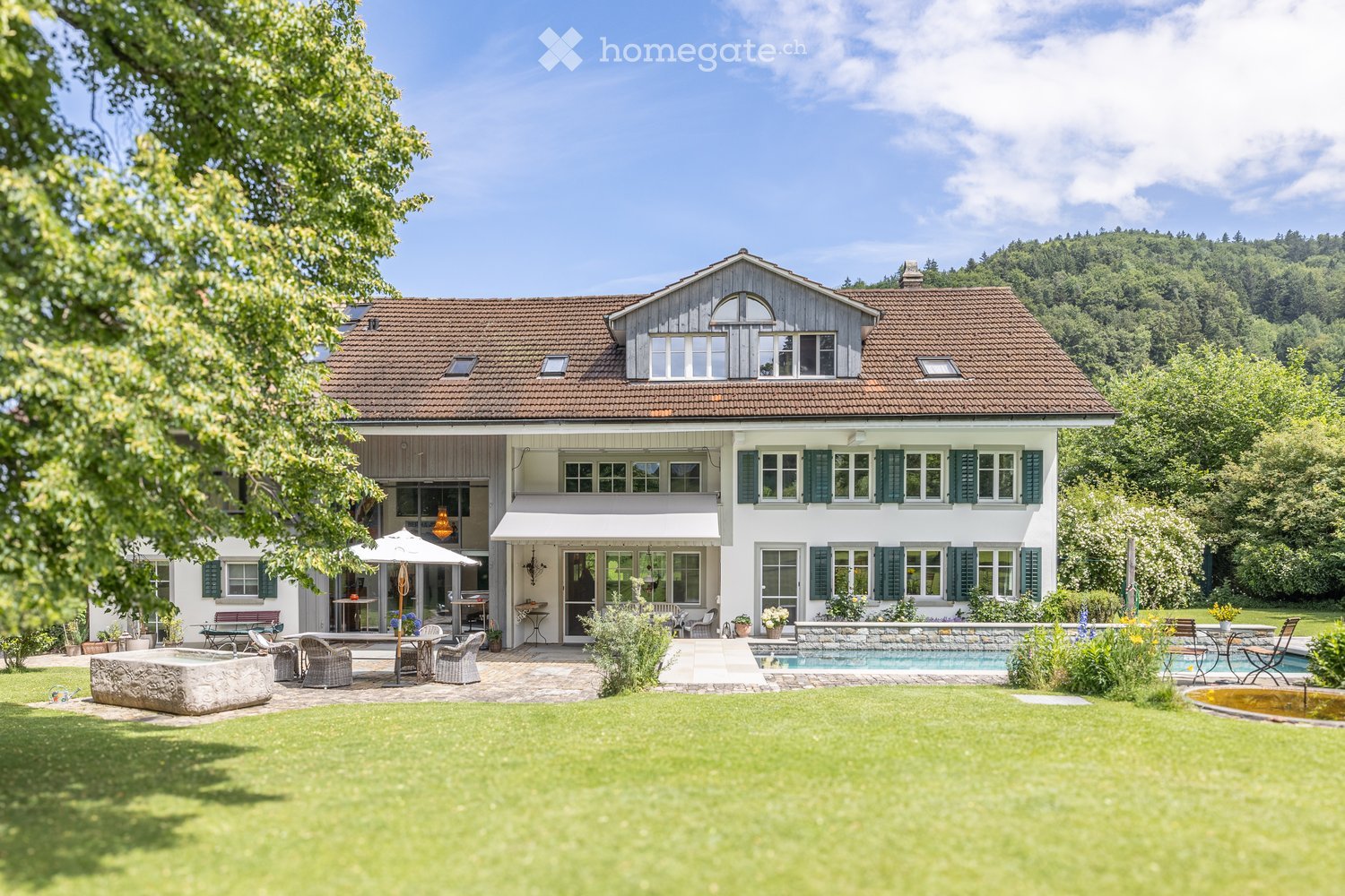 Two-story house with a tiled roof, white walls, green shutters, and a spacious garden with a swimming pool, outdoor seating, and a patio area.