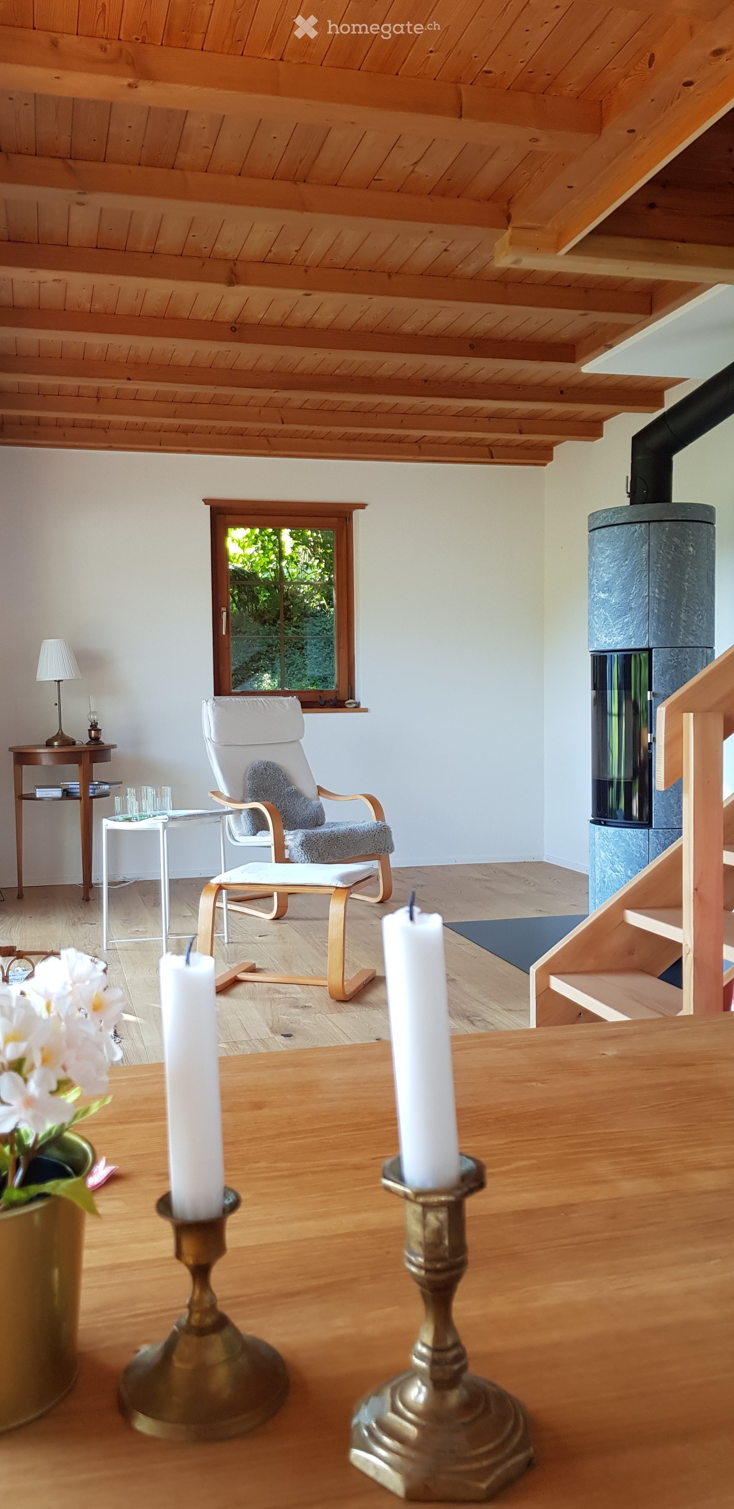 Bright living room with wooden floors, white walls, and exposed wooden ceiling beams. Features a white armchair with a grey cushion, a small wooden side table with a lamp, and two tall candles on the floor.