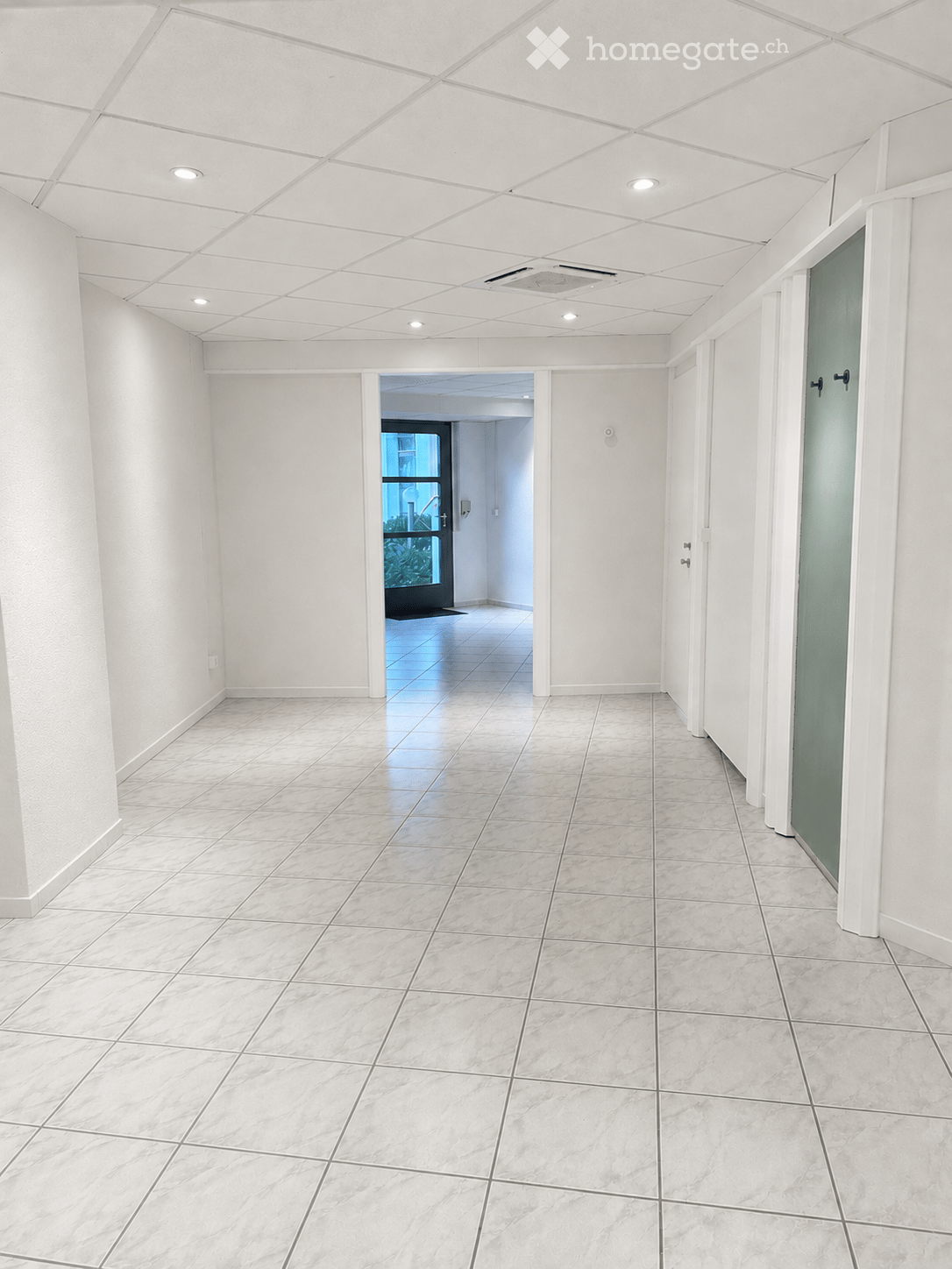 Empty hallway with white walls, tiled floor, and recessed lighting. Features a door leading to another room and a glass door with a view of greenery outside.