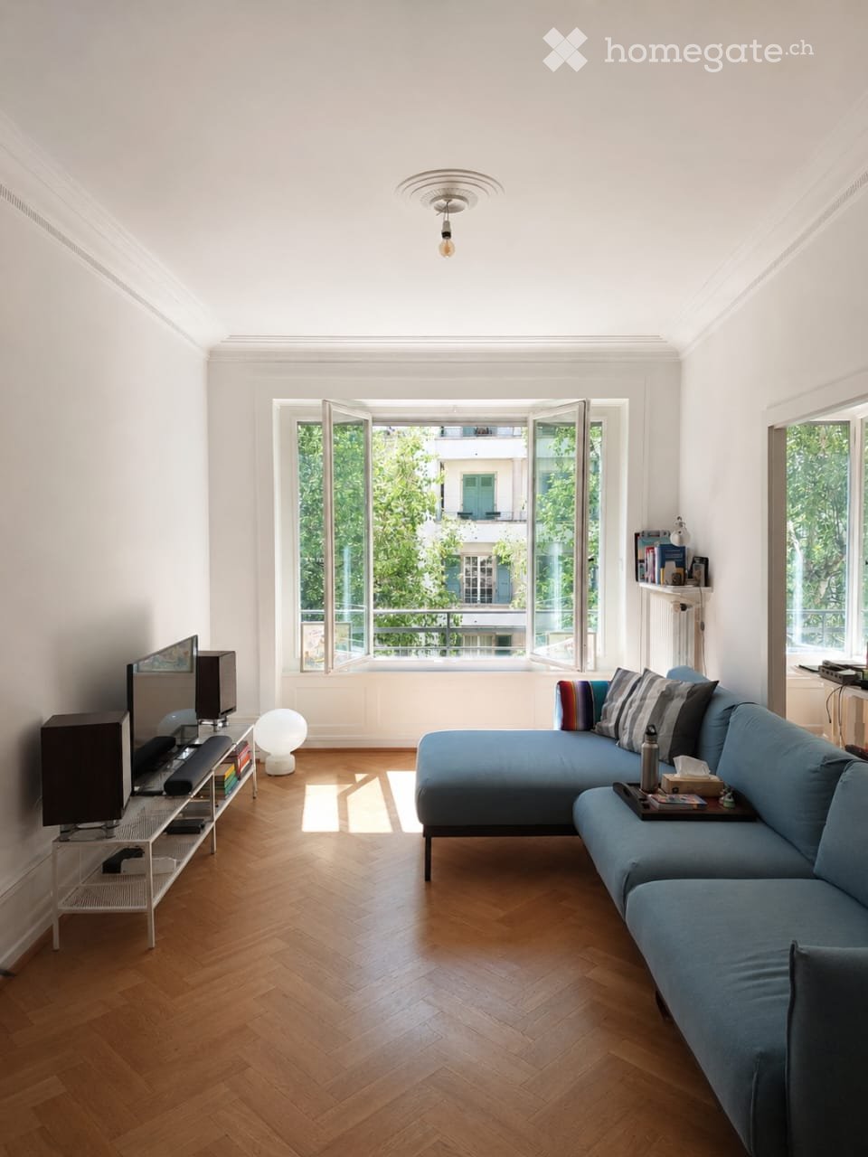 Living room with wooden floor, large windows, blue sofa, white ball lamp, TV, and bookshelf.