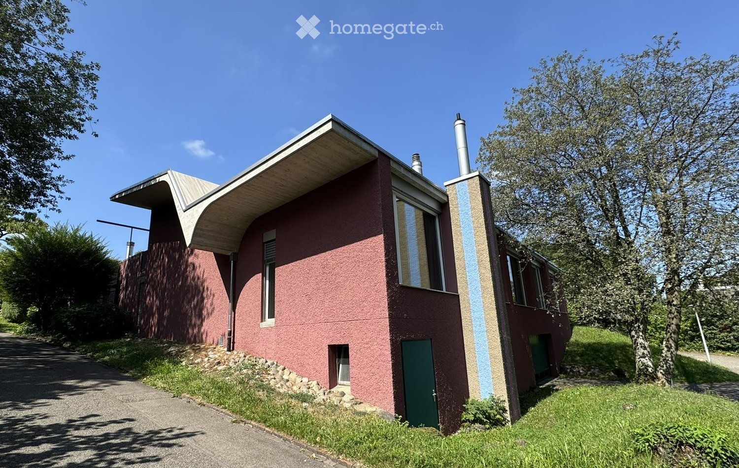 Modern single-story house with a distinctive curved roof and red textured facade, featuring large windows and a green door.
