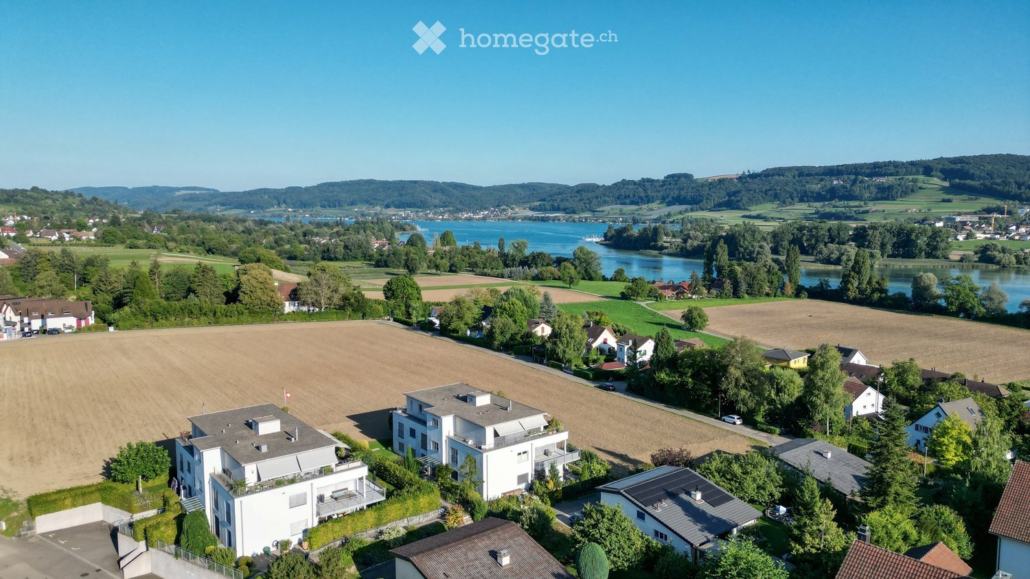 Aerial view of a suburban neighborhood with multiple houses, green fields, and a body of water in the background.