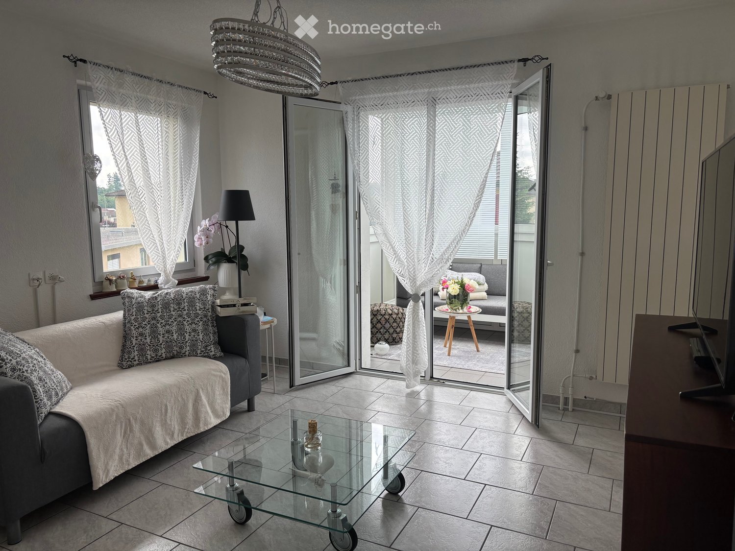 Modern living room with a grey sofa, glass coffee table, floor lamp, and a view of a balcony area with a small seating set.