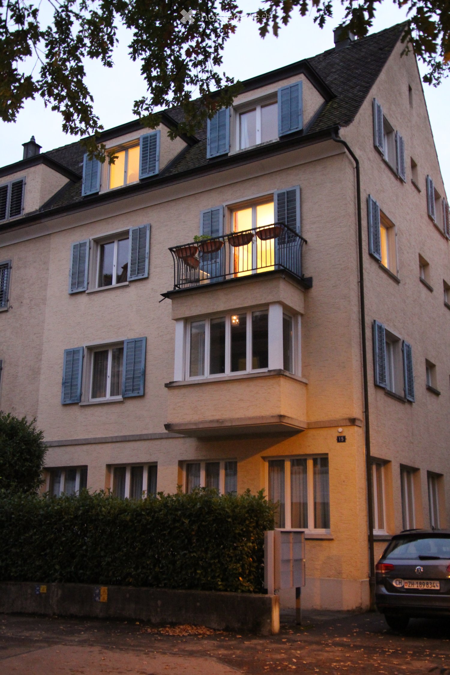 A multi-story residential building with light-colored walls, multiple windows with shutters, and a balcony with potted plants. The building has a classic architectural style with a pitched roof and visible lighting inside.