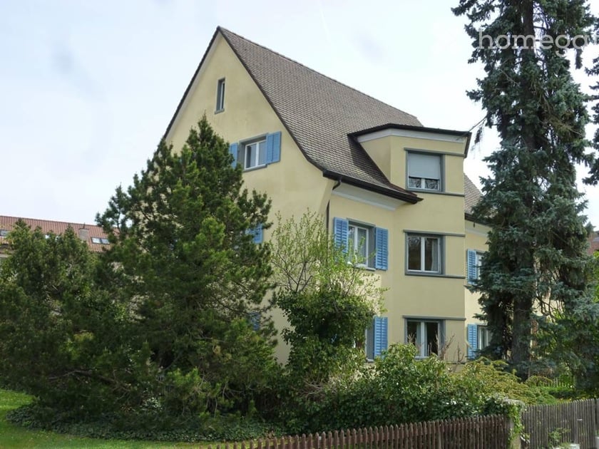 Two-story yellow house with blue shutters, surrounded by trees and a wooden fence.