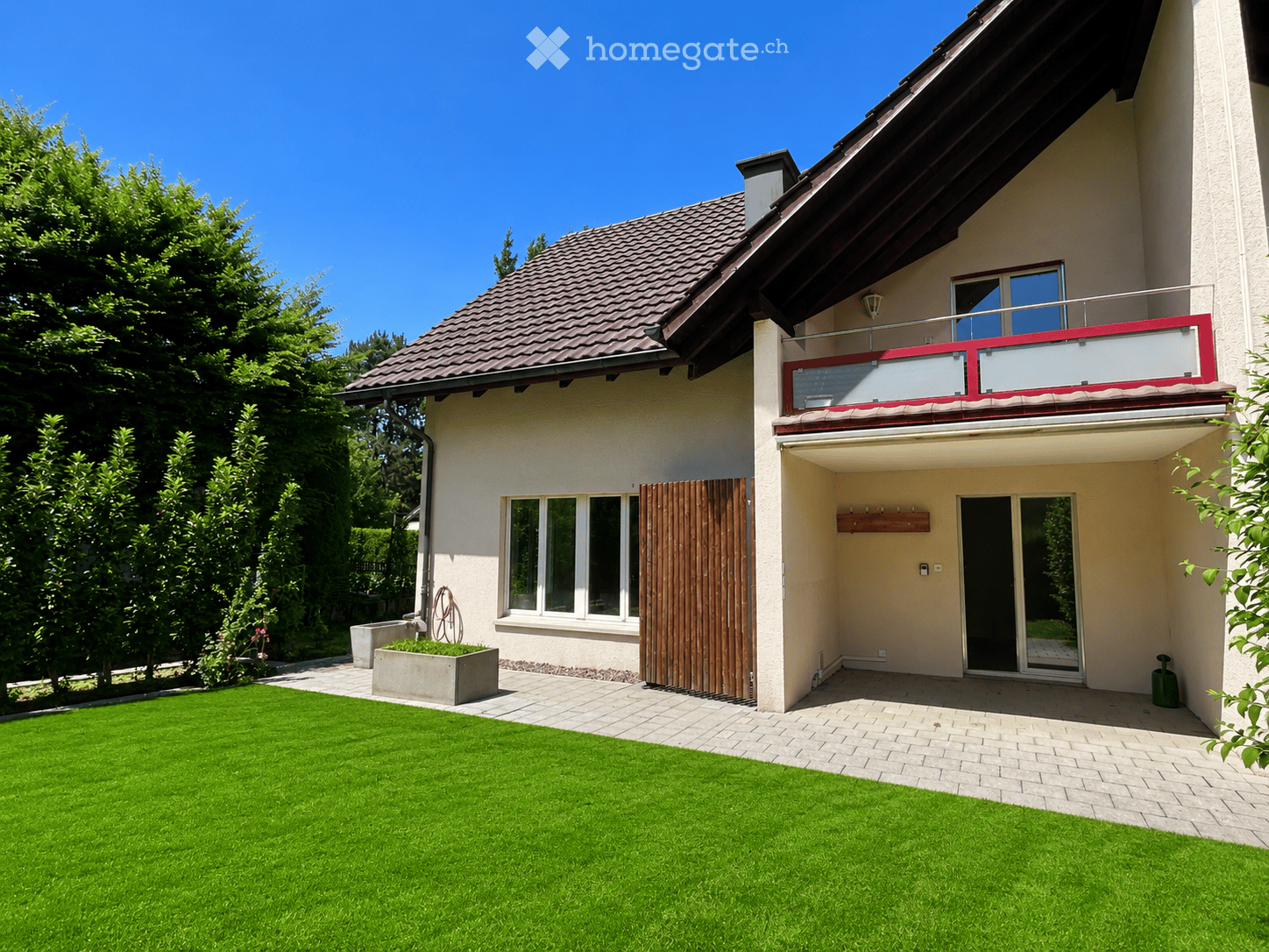 Two-story house with a red-tiled roof, white walls, large windows, and a balcony on the upper floor. The front yard features a well-maintained lawn, a brick pathway, and a small planter with greenery.