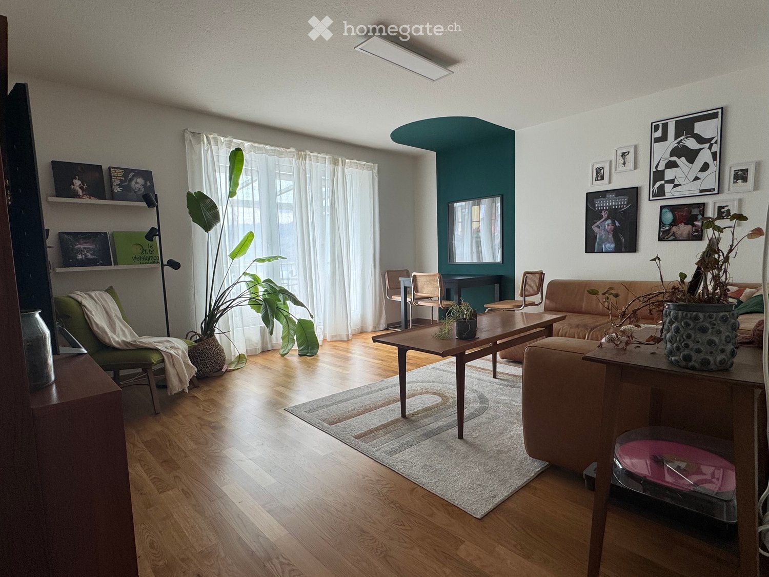 Modern living room with wooden flooring, a green accent wall, large windows with white curtains, a beige sofa, a wooden coffee table, a green armchair, and decorative shelves with artwork.