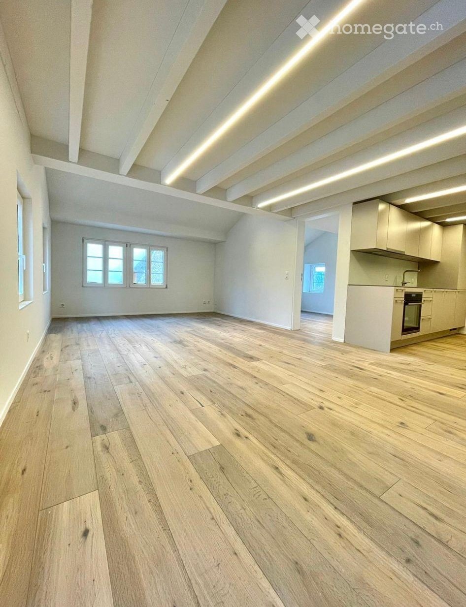 Empty room with wooden flooring, white walls, and exposed wooden beams on the ceiling. The room has large windows and an open doorway leading to a small kitchen area.