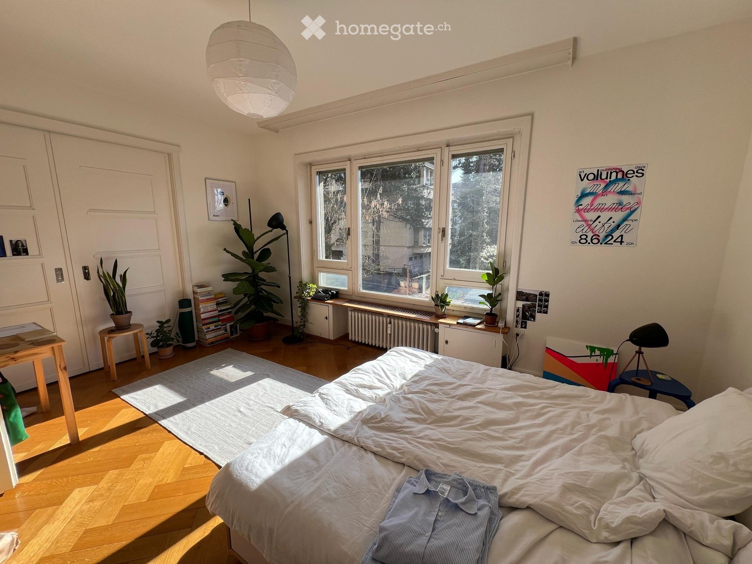 Cozy bedroom with a large bed, wooden floor, large windows, and various decorative items including plants, a lamp, and artwork.