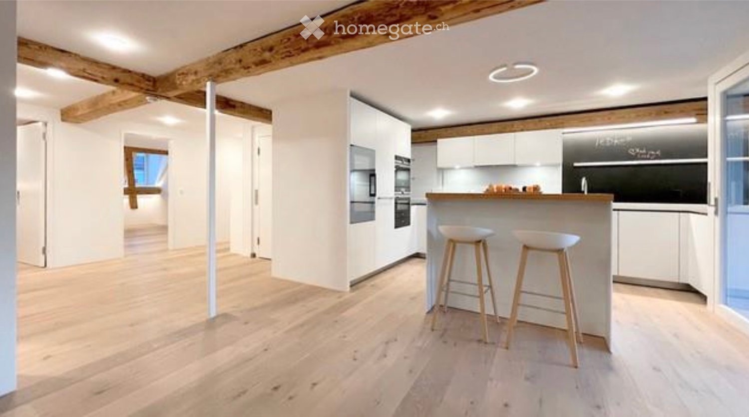 Modern kitchen with white cabinets, wooden bar counter, two stools, and built-in appliances including an oven and refrigerator.