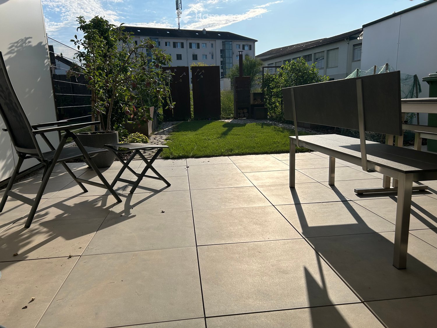 A modern patio with a tiled floor, a folding chair, a small table, and a bench, surrounded by greenery and a well-maintained lawn.