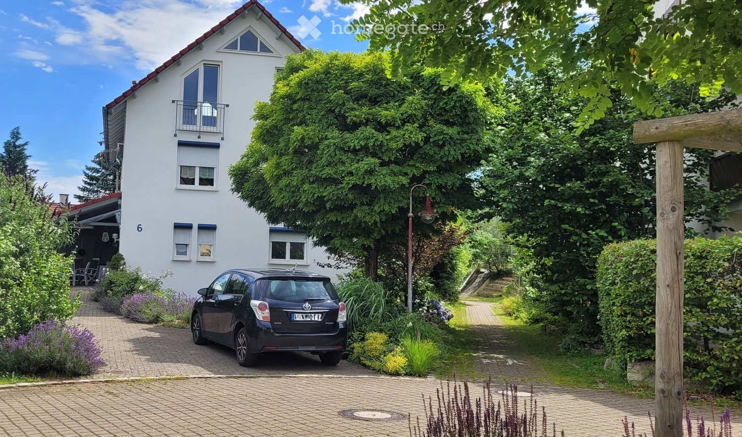 Two-story white house with a red-tiled roof, multiple windows, and a balcony on the upper floor. A small black car is parked on a paved driveway surrounded by lush greenery and flowering plants.