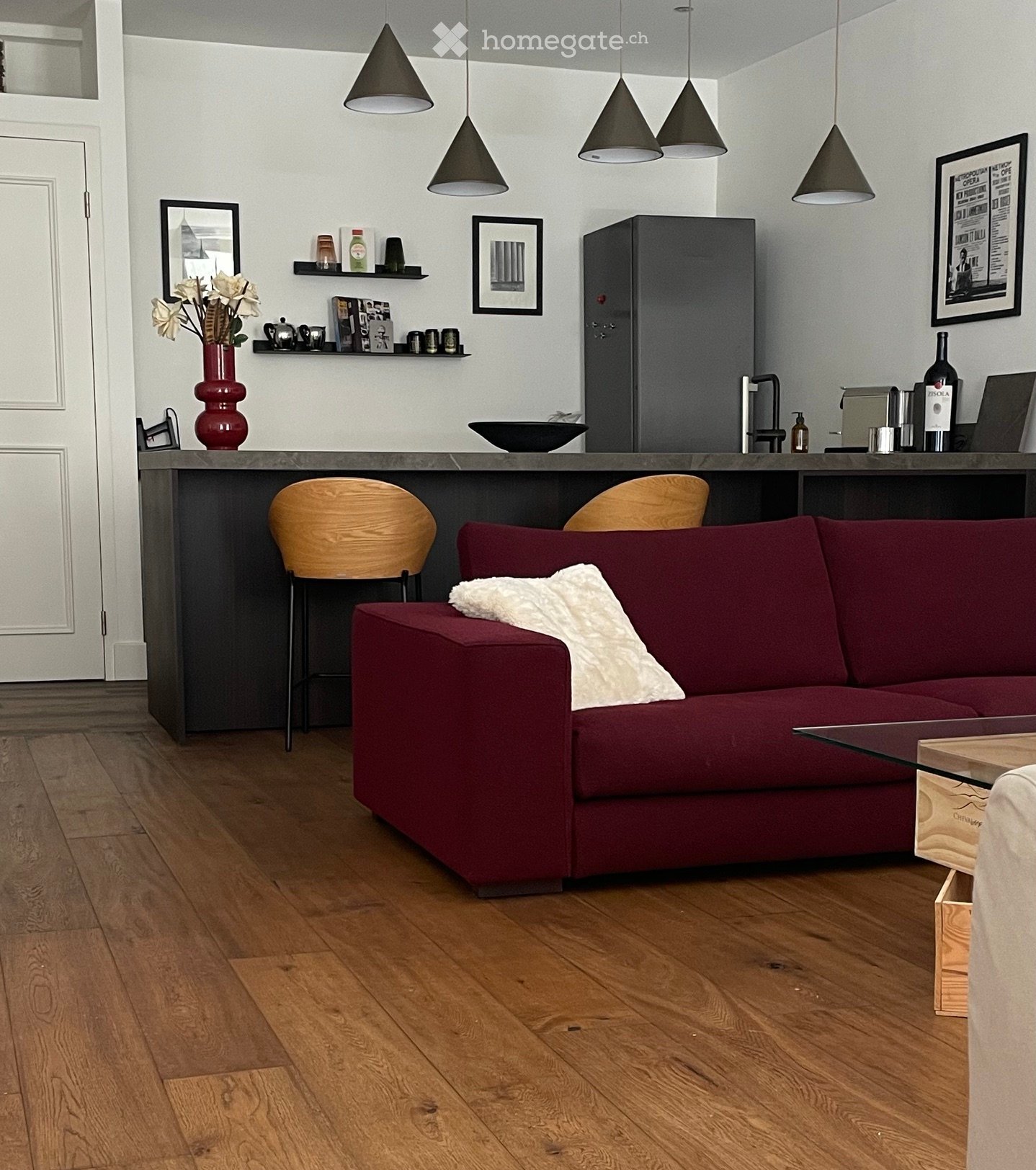 Modern living room with a red sofa, coffee table, wooden floor, pendant lights, and a bar area with a refrigerator and stools.