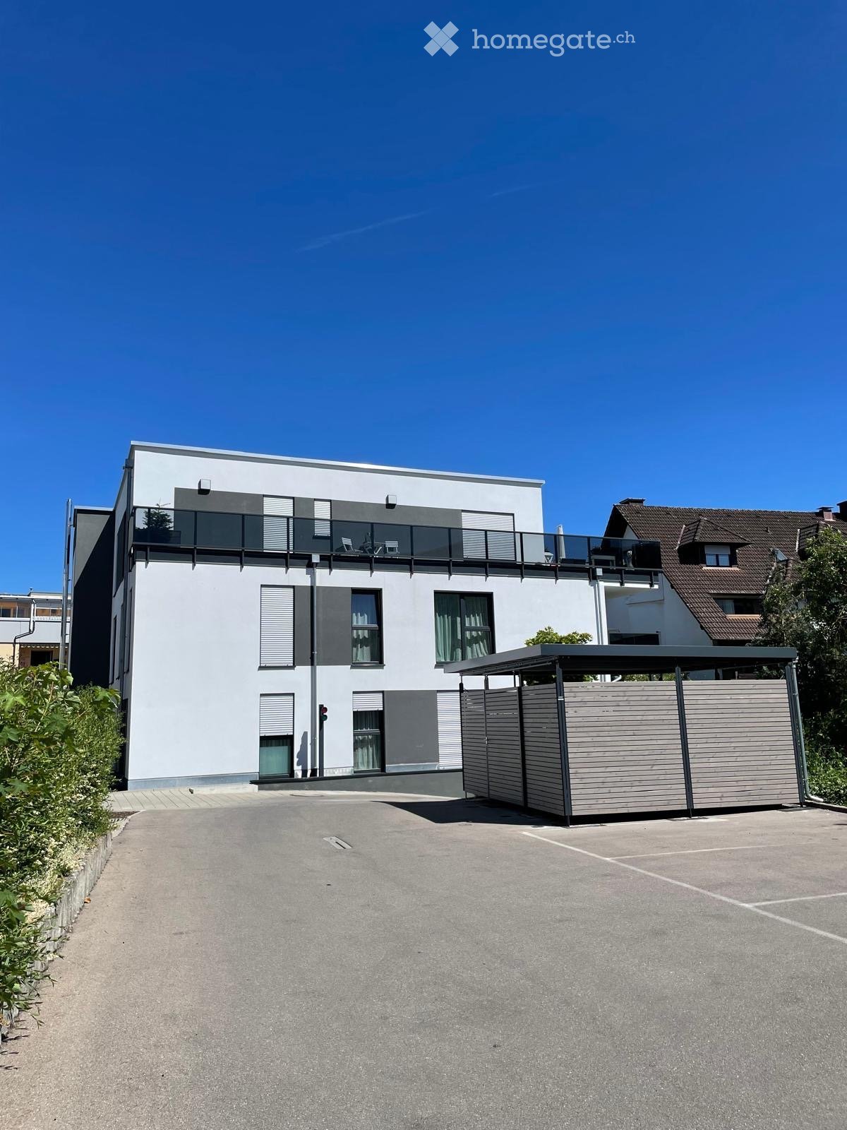 Modern two-story house with white facade, large windows, and a rooftop terrace with railings. Features a covered parking area and a landscaped driveway.