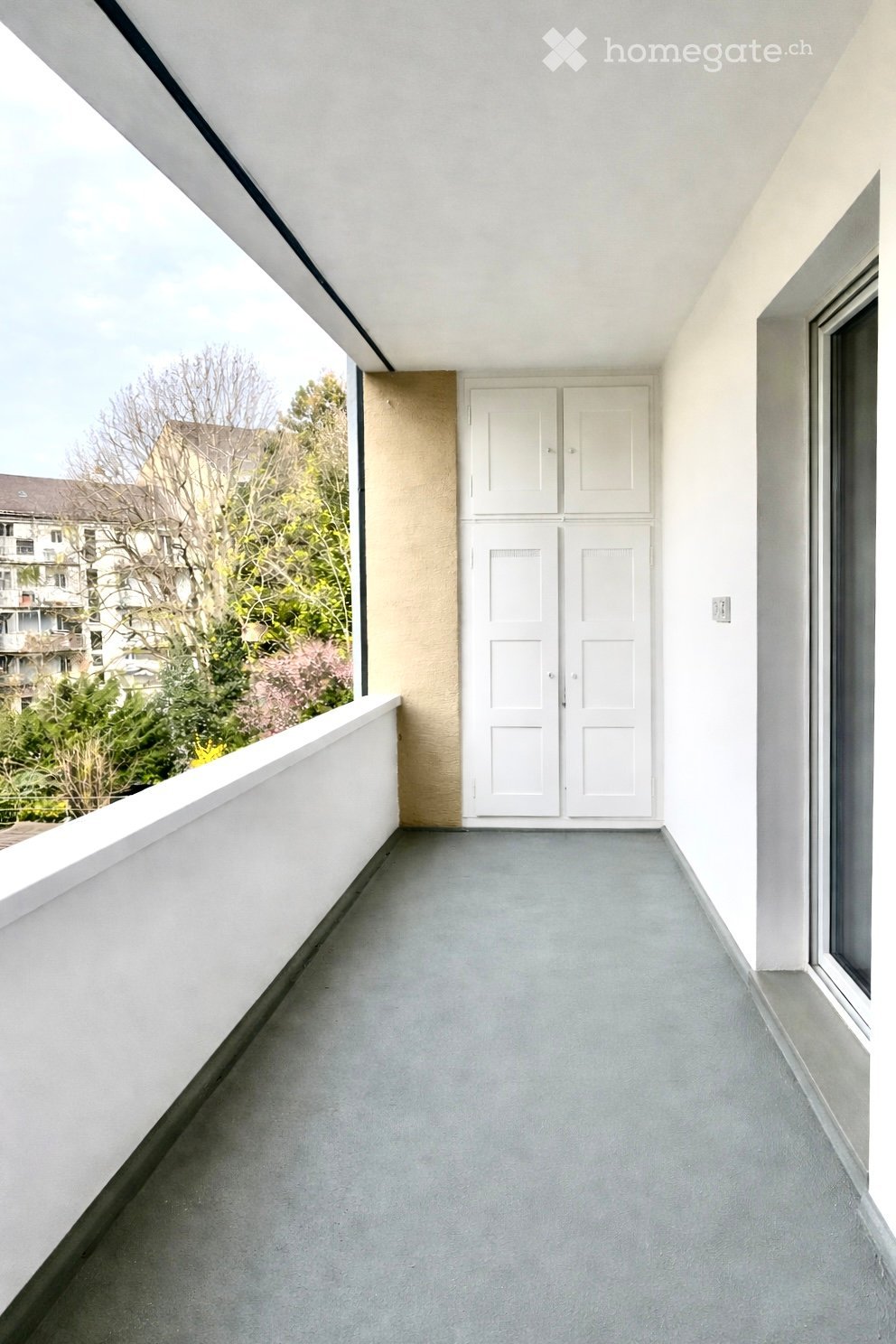Modern balcony with white walls, grey floor, large window, and double doors leading inside.