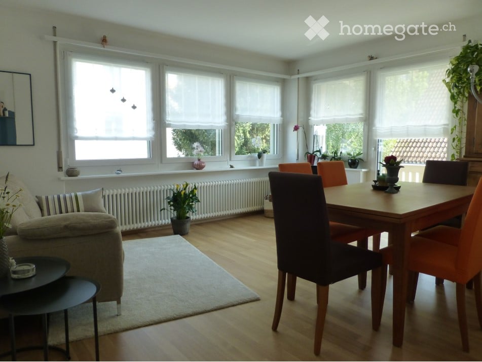 Bright living and dining area with large windows, wooden flooring, a beige sofa, a coffee table, a dining table with chairs, and several potted plants.