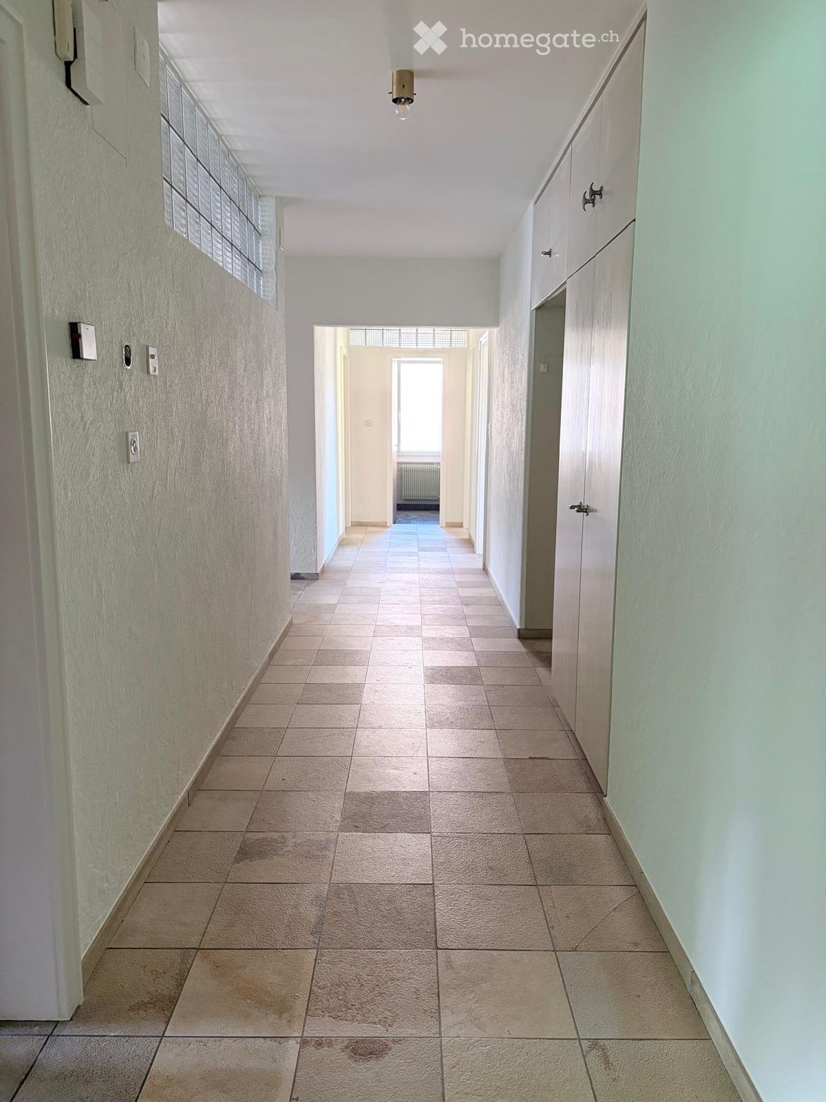 Empty hallway with tiled floor, light-colored walls, and a single ceiling light fixture. The hallway leads to an open area with a window at the end.