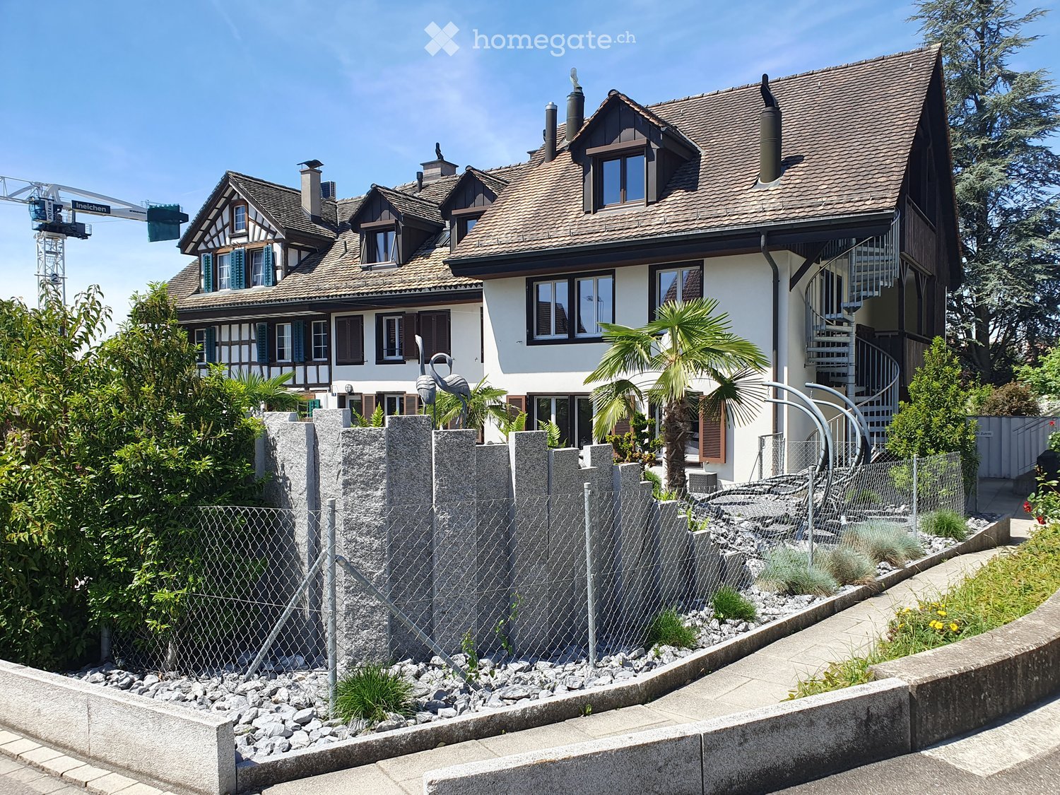 Two-story house with a tiled roof, multiple windows, and a decorative garden area with a palm tree and a flamingo sculpture.