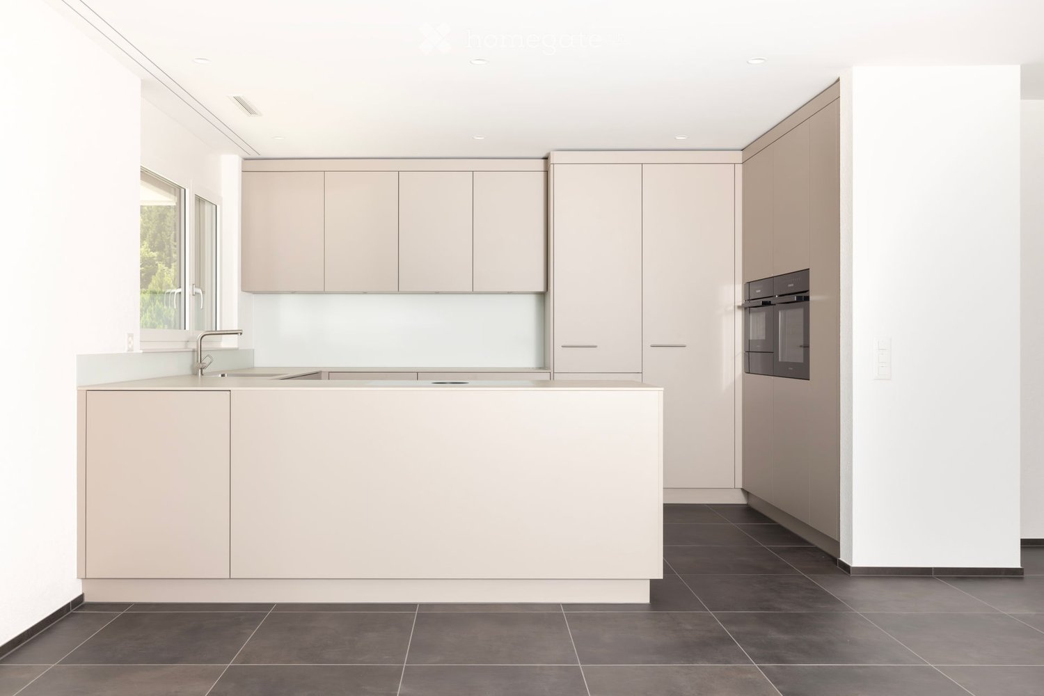Modern kitchen with light-colored cabinets, a central island, and dark tiled flooring. Features a built-in oven and a sleek sink with a modern faucet.