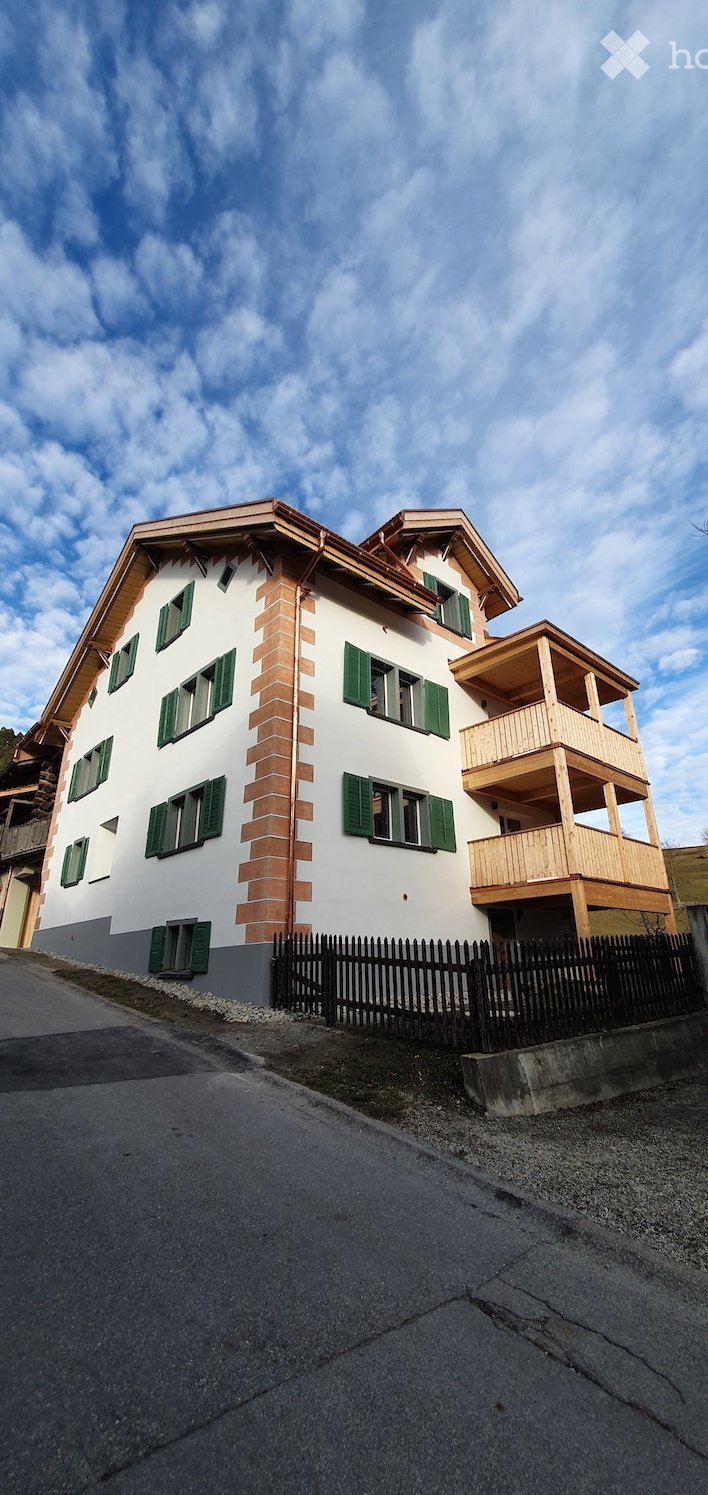 Two-story house with white walls, green shutters, and a wooden balcony on the upper level.