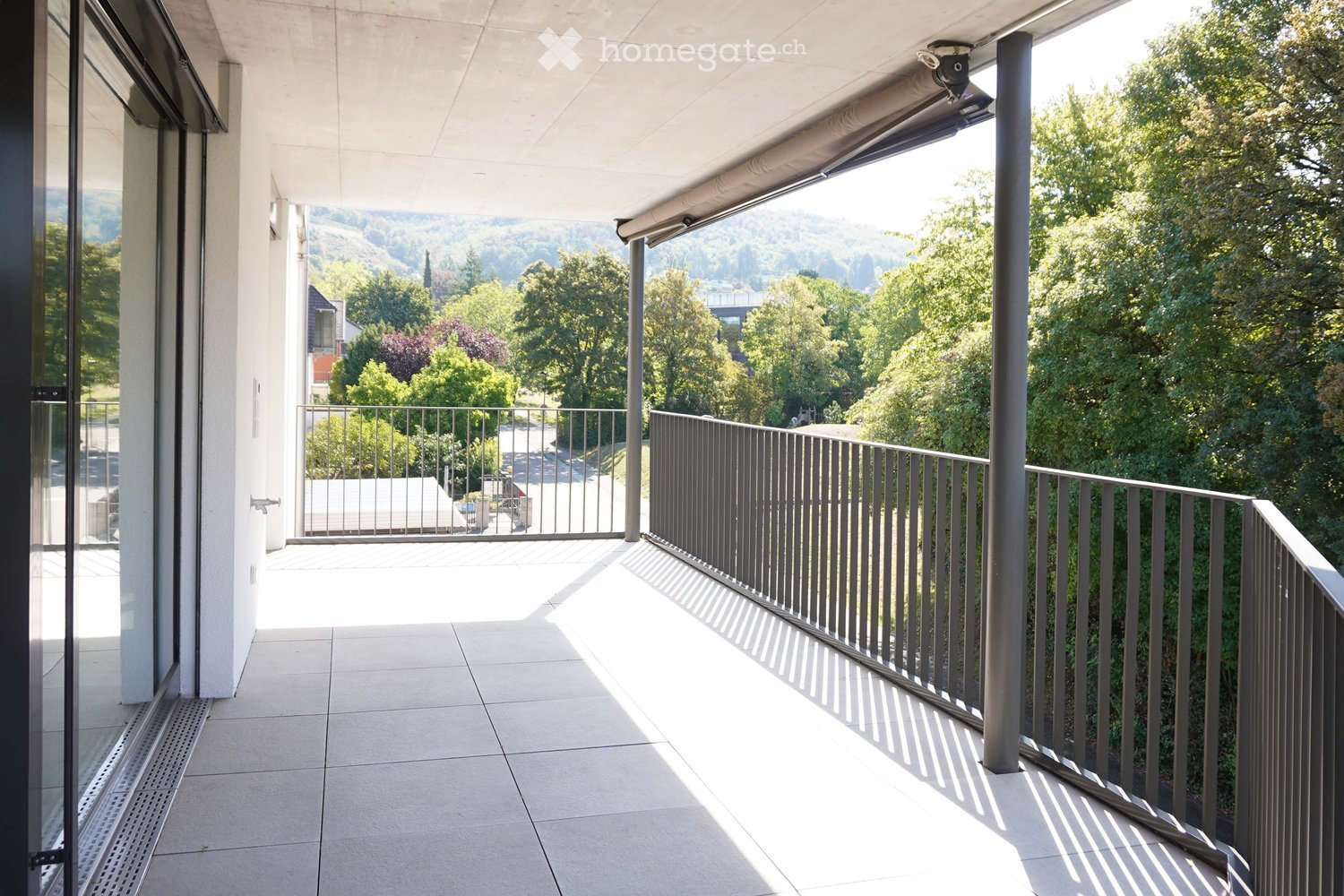 Balcony with tiled floor, metal railing, and retractable awning, overlooking a green, tree-filled landscape.