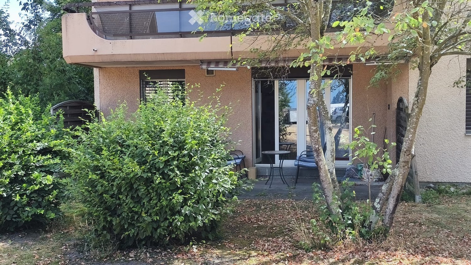 A small patio area with a table and chairs, surrounded by greenery and a tree, attached to a two-story building with a balcony above.