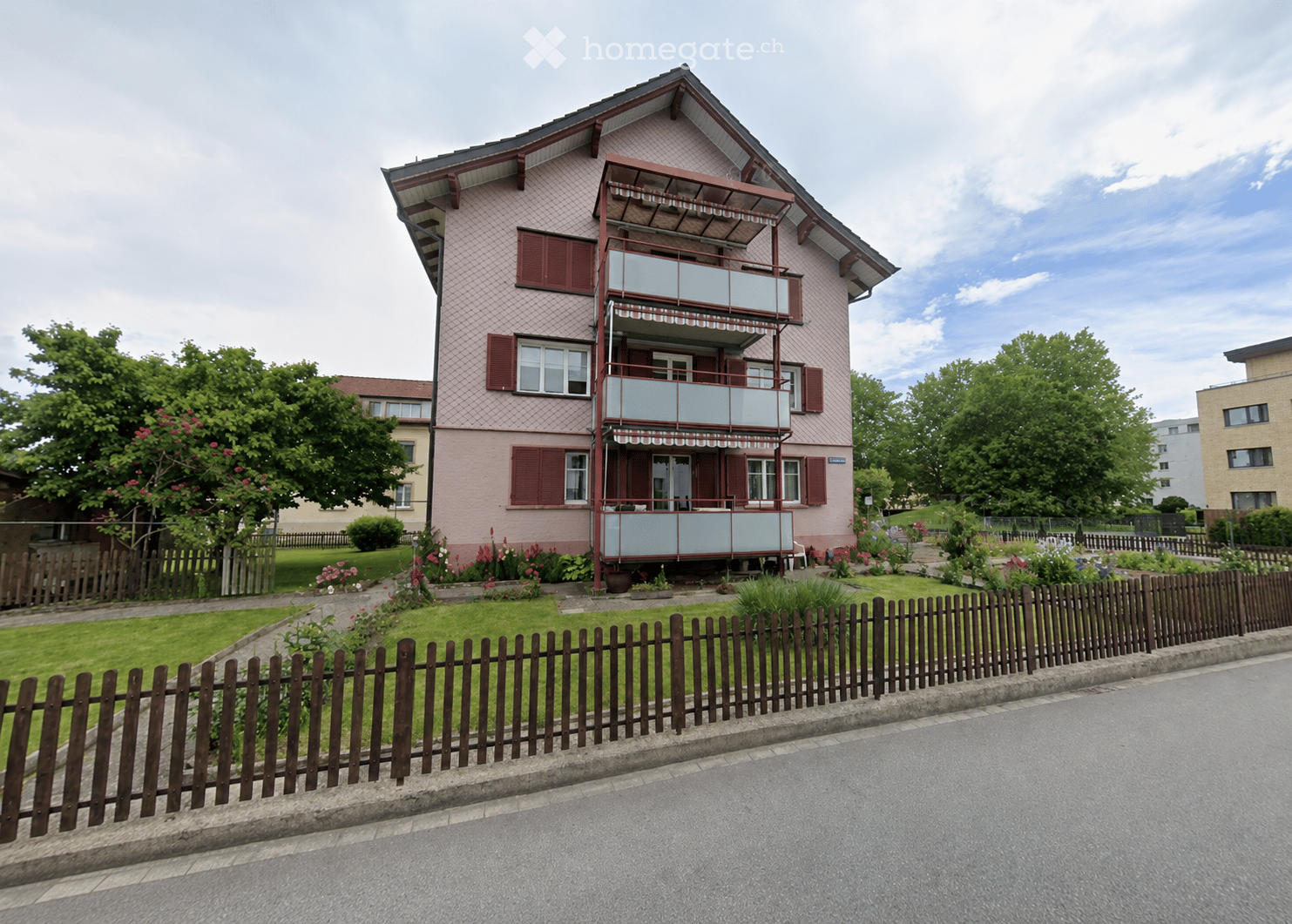 Two-story house with pink exterior, red shutters, and a balcony with glass railing. The house is surrounded by a wooden fence, garden, and trees.
