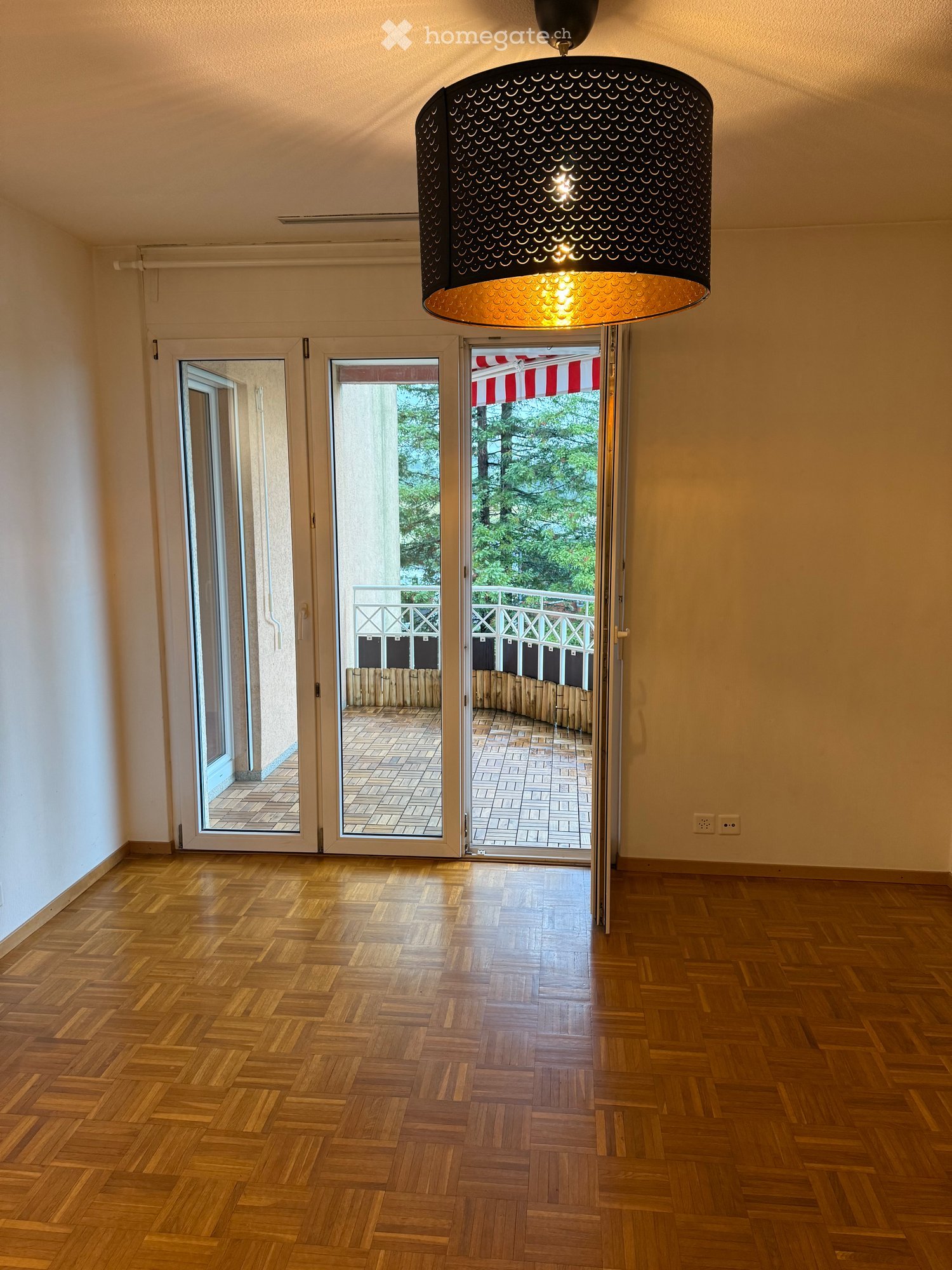 Empty hallway with wooden flooring, large pendant light fixture, and three sets of glass doors leading to a balcony area with tiled flooring and a striped awning visible outside.
