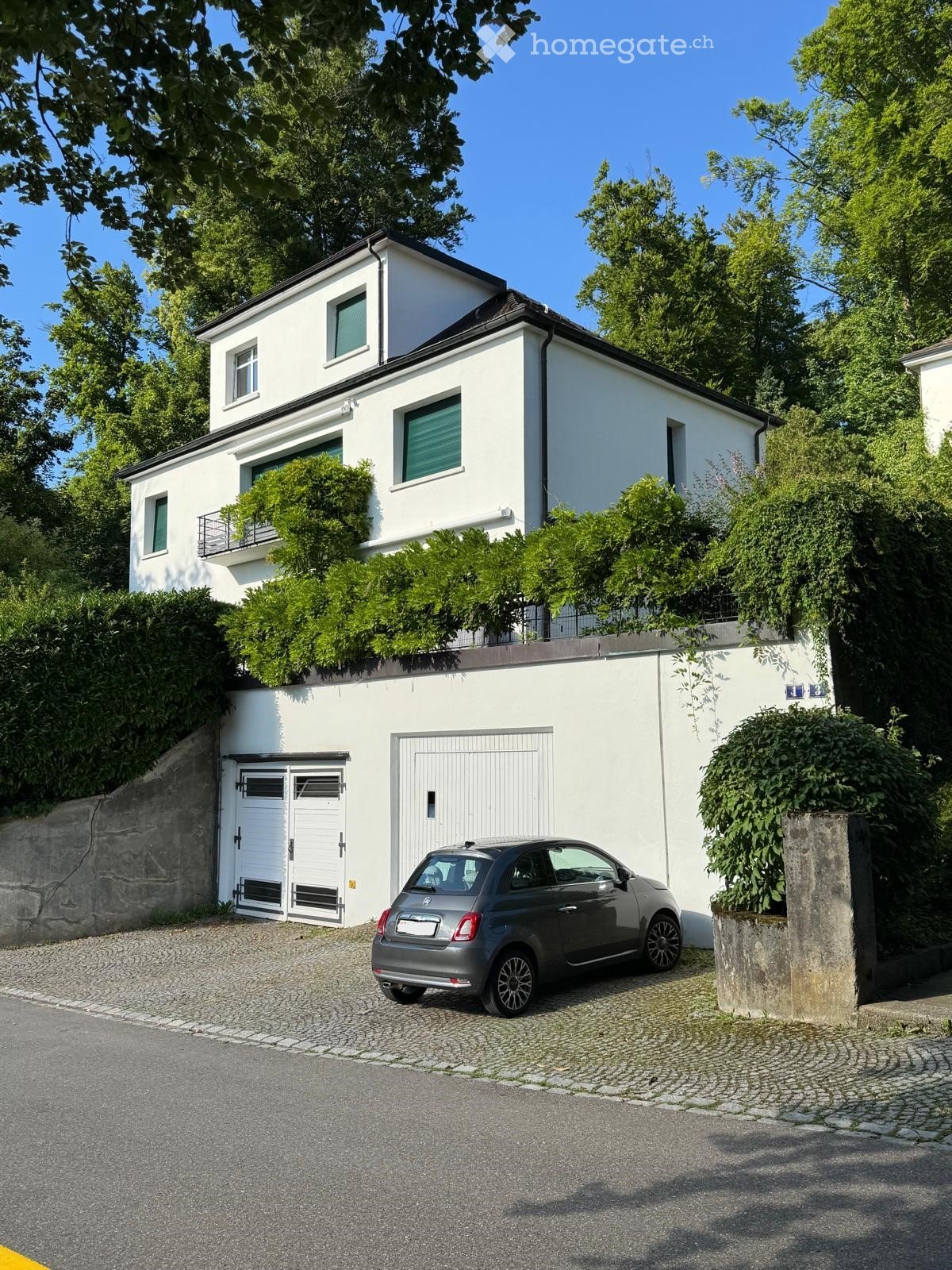 Two-story white house with multiple windows, a garage, and a small balcony on the upper level. A car is parked in front of the garage.