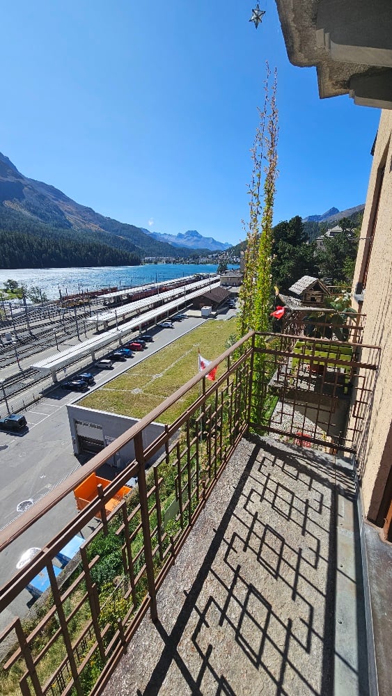 Balcony with metal railing overlooking a scenic view of mountains, a river, and a town below. The area includes a small garden patch and a few parked cars in the distance.