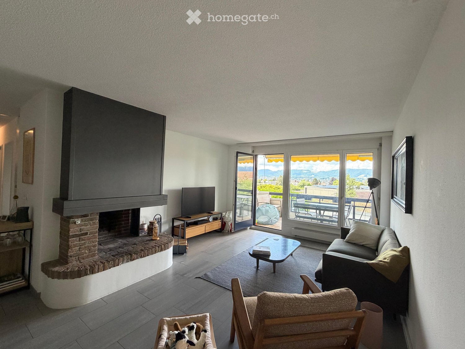 Bright living room with large windows offering a scenic view, a fireplace, a sofa, coffee table, armchair, and a television.
