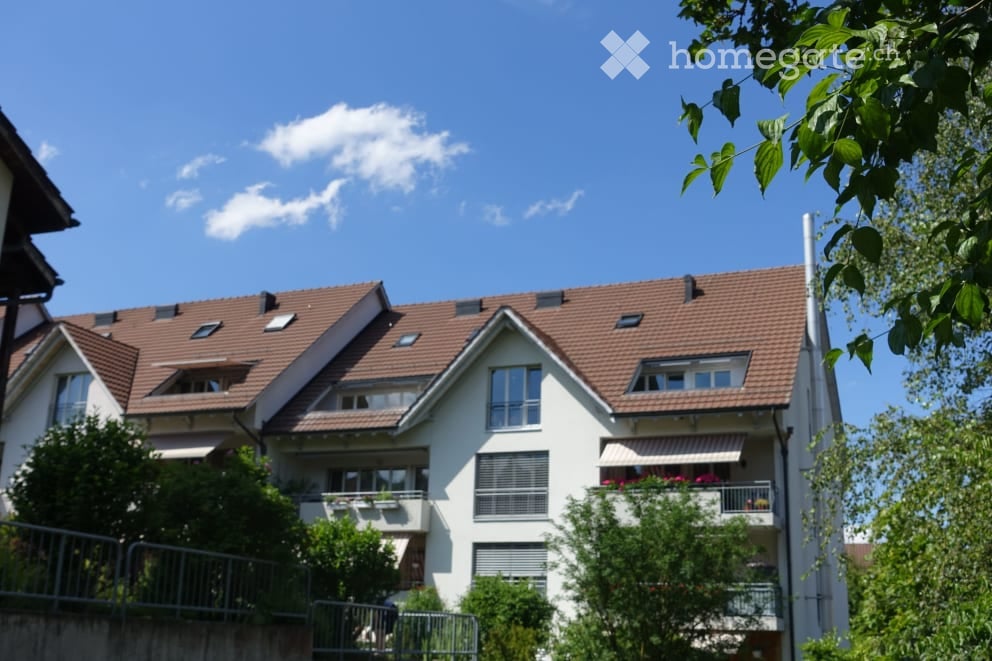 A modern two-story residential building with a red-tiled roof, white exterior walls, multiple windows, and balconies adorned with plants and flowers.