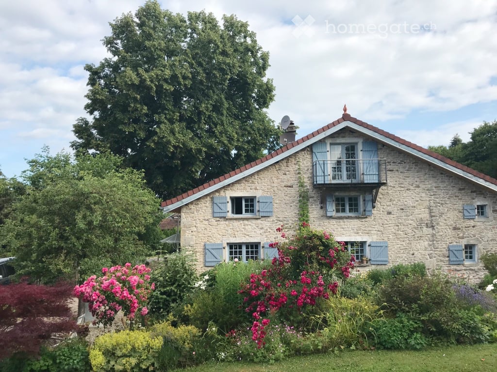 Stone house with blue shutters, balcony, surrounded by lush garden and trees.