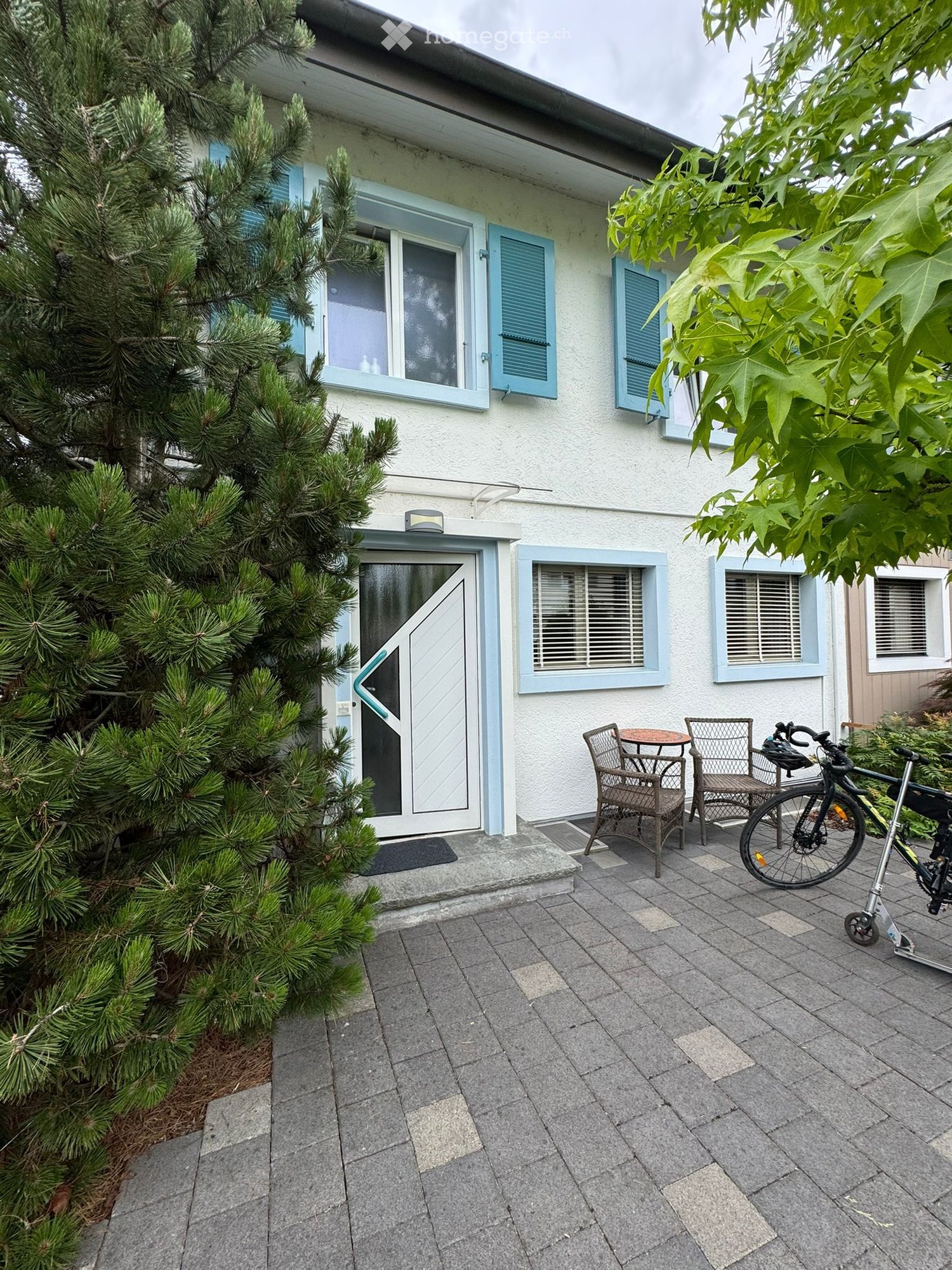 Paved outdoor area with a small seating area, including two chairs and a table, a bicycle, and a tree near the entrance of a house.