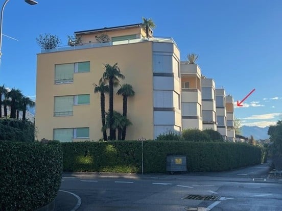 Modern multi-story apartment building with light-colored facade, large windows, and palm trees in front. The building has multiple balconies and a rooftop terrace.