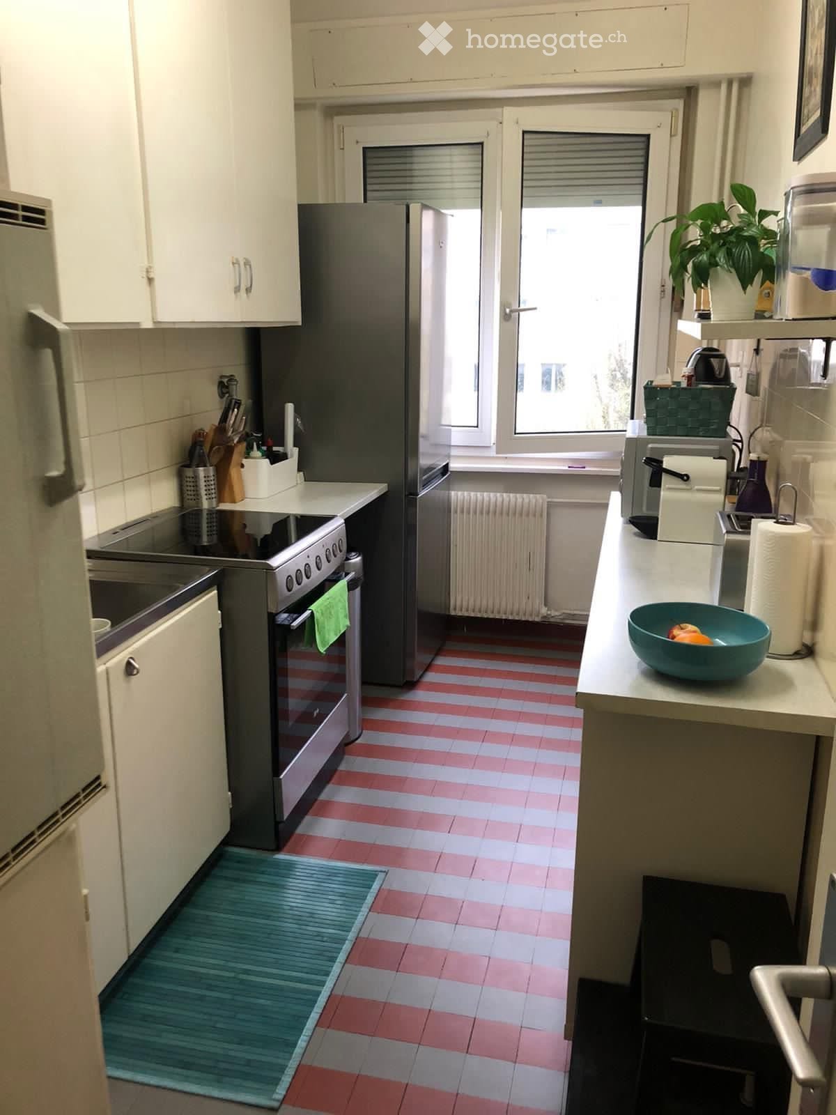 A compact kitchen with white cabinets, a stainless steel refrigerator, an oven, a sink, and a countertop with a bowl of fruit. The floor features a red and white tiled pattern.
