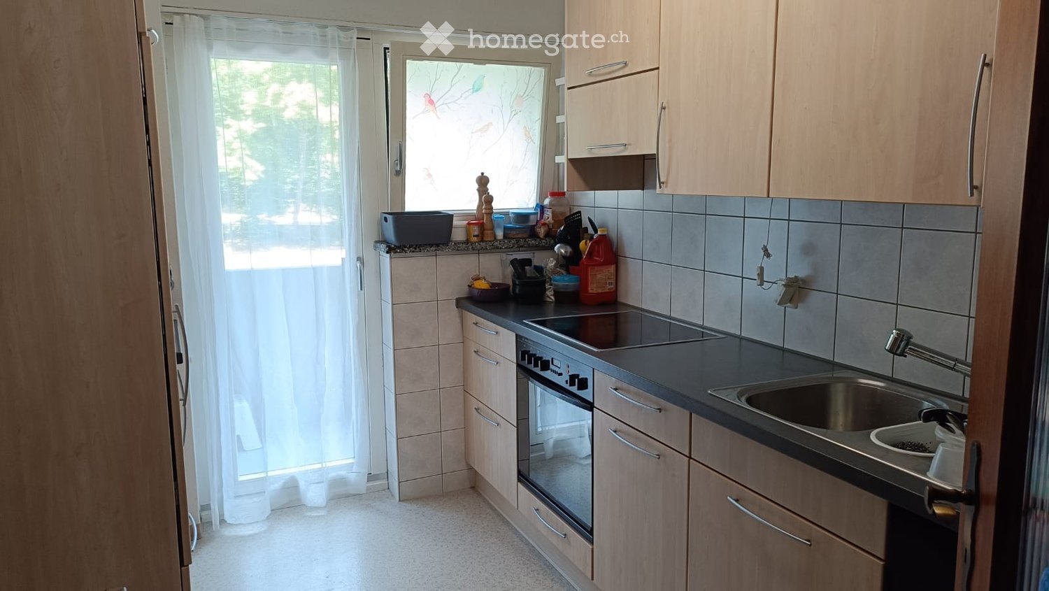 Modern kitchen with light wooden cabinets, stainless steel sink, electric stove, and various small appliances and items on the counter.