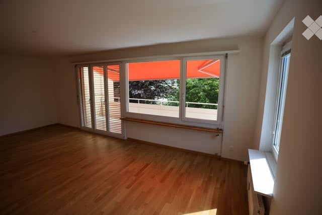 Empty room with wooden flooring, large windows, and sliding glass doors leading to a balcony.