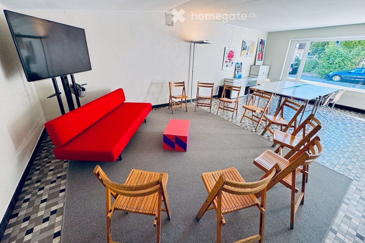 A modern, open room with a red sofa, multiple wooden chairs, a large screen, and a table with chairs arranged for meetings or presentations.