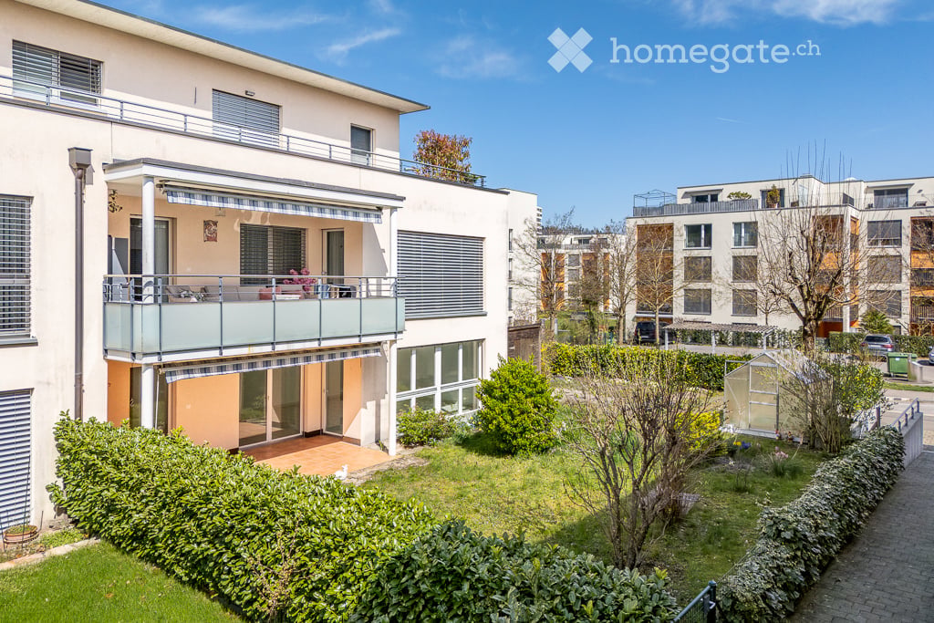 Modern multi-story residential building with balconies, surrounded by a well-maintained garden and neighboring structures.