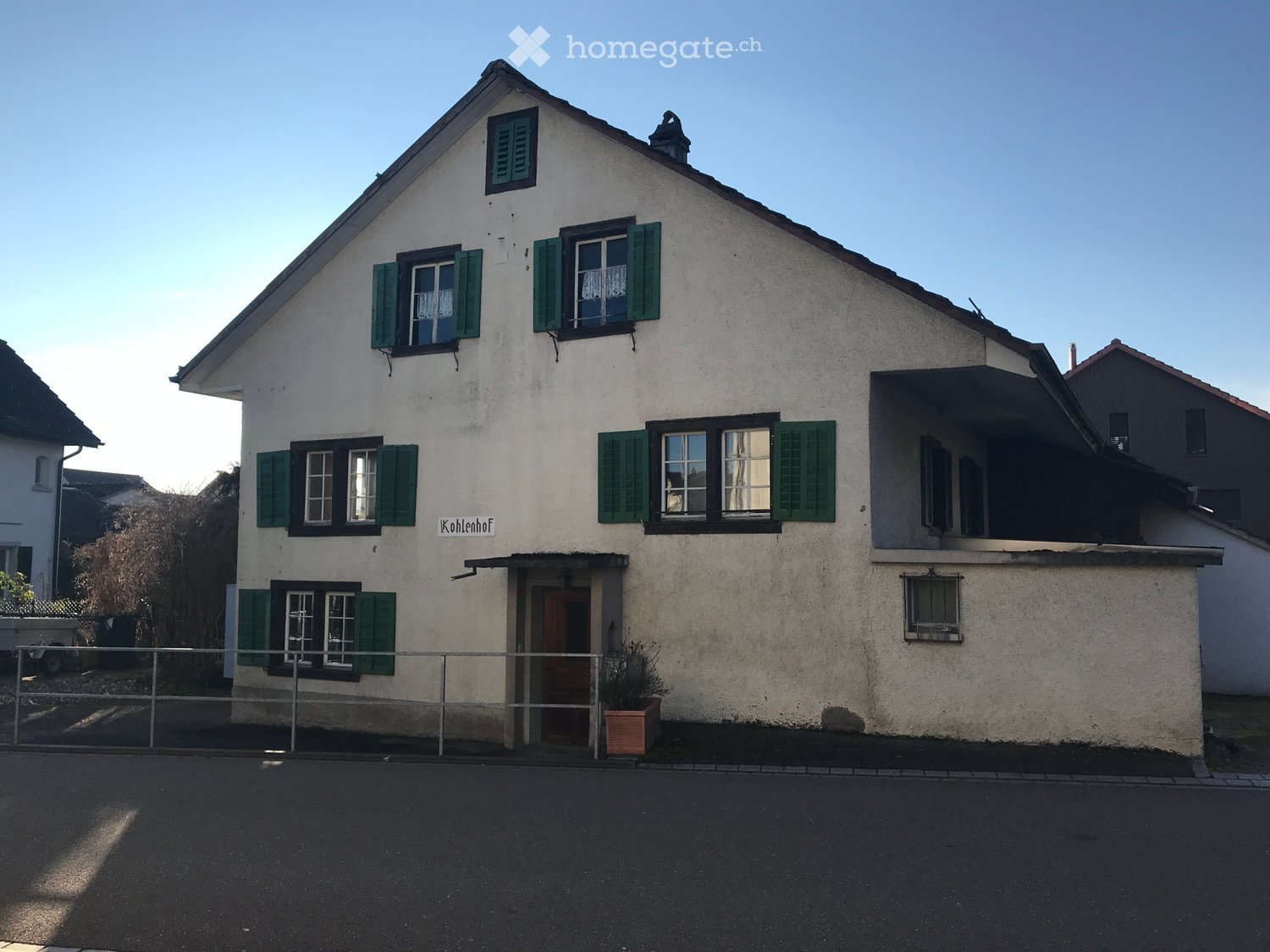Two-story house with white plaster walls, green shutters, and a pitched roof. Features a covered entrance porch and a small balcony on the upper floor.
