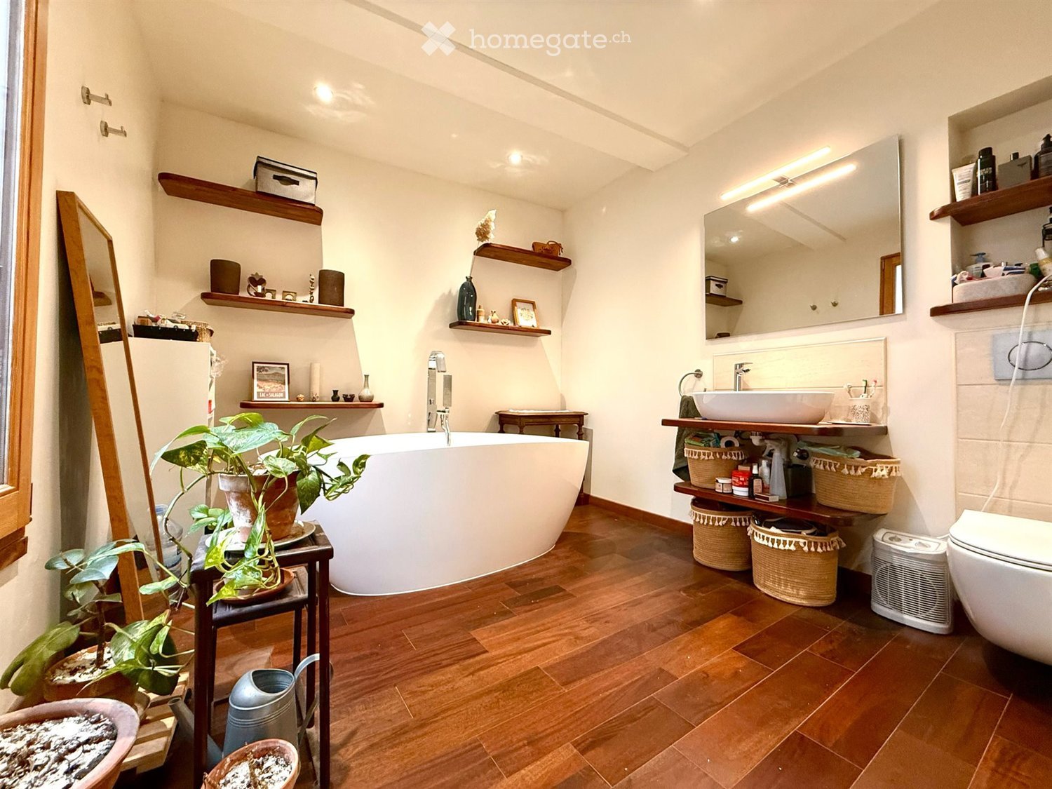 Bathroom with wooden flooring, large bathtub, double sink, toilet, mirror, wooden shelves, decorative items, and plants.