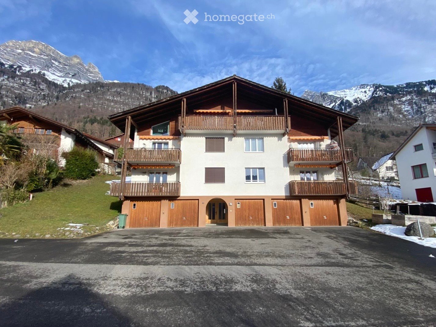 3-story building, brown wooden balconies, white and brown walls, wooden garage doors, mountain backdrop