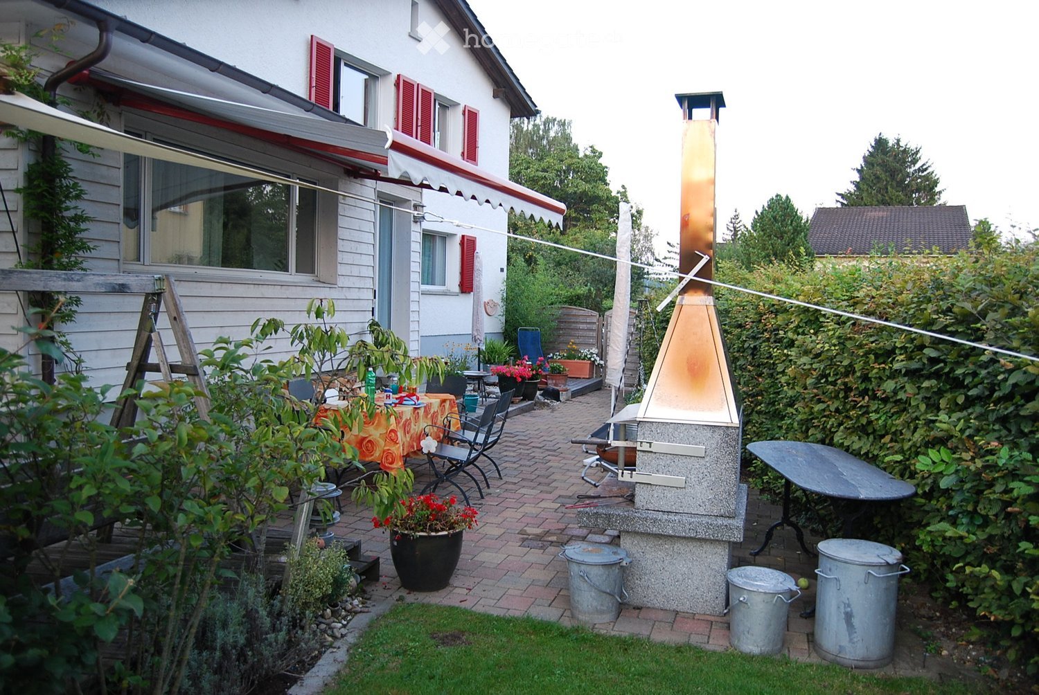 A well-maintained garden area with a brick patio, outdoor dining table, chairs, barbecue, potted plants, and a small grassy lawn.