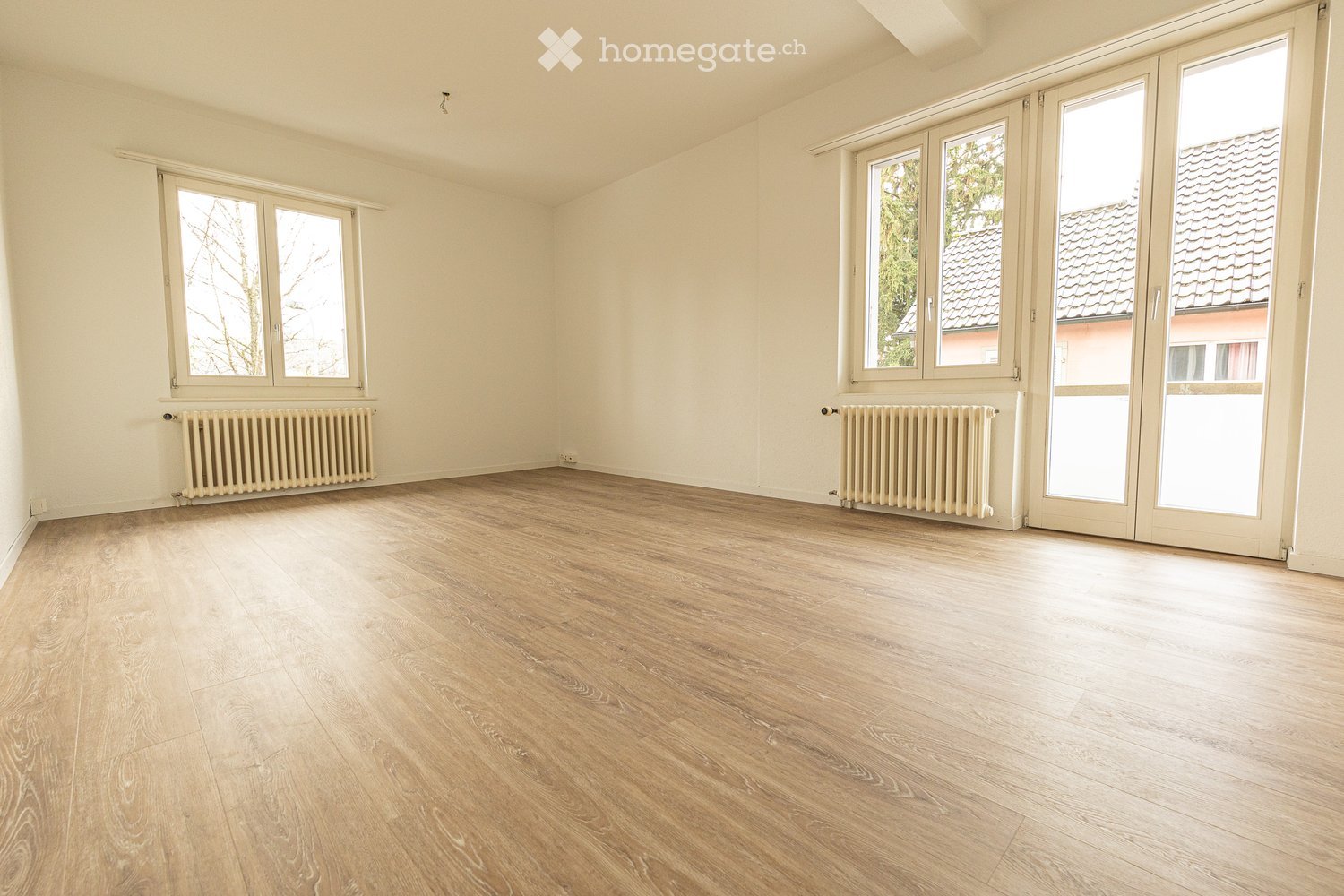 Empty room with light wooden flooring, white walls, and large windows with radiators beneath them.