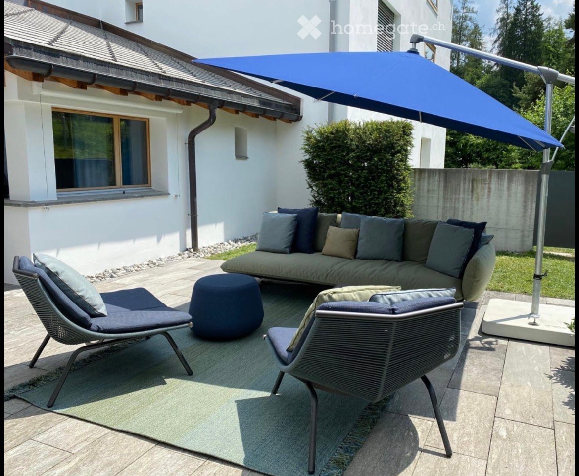 Outdoor patio with a grey sectional sofa, two wicker chairs, an ottoman, and a large blue umbrella providing shade. The area is surrounded by a white building and greenery.