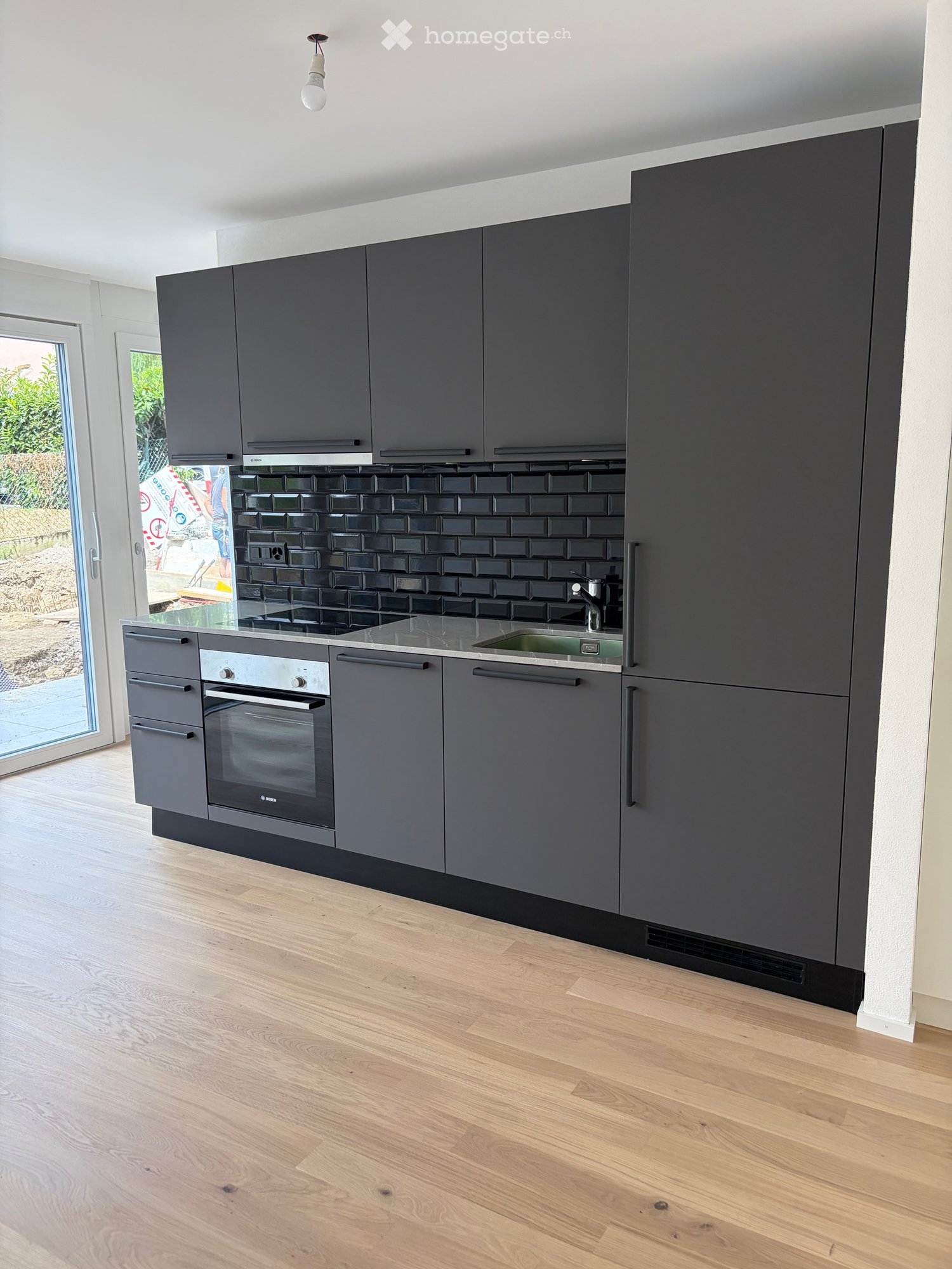Modern kitchen with grey cabinets, stainless steel oven, sink, and faucet, featuring a black tiled backsplash and wooden flooring.